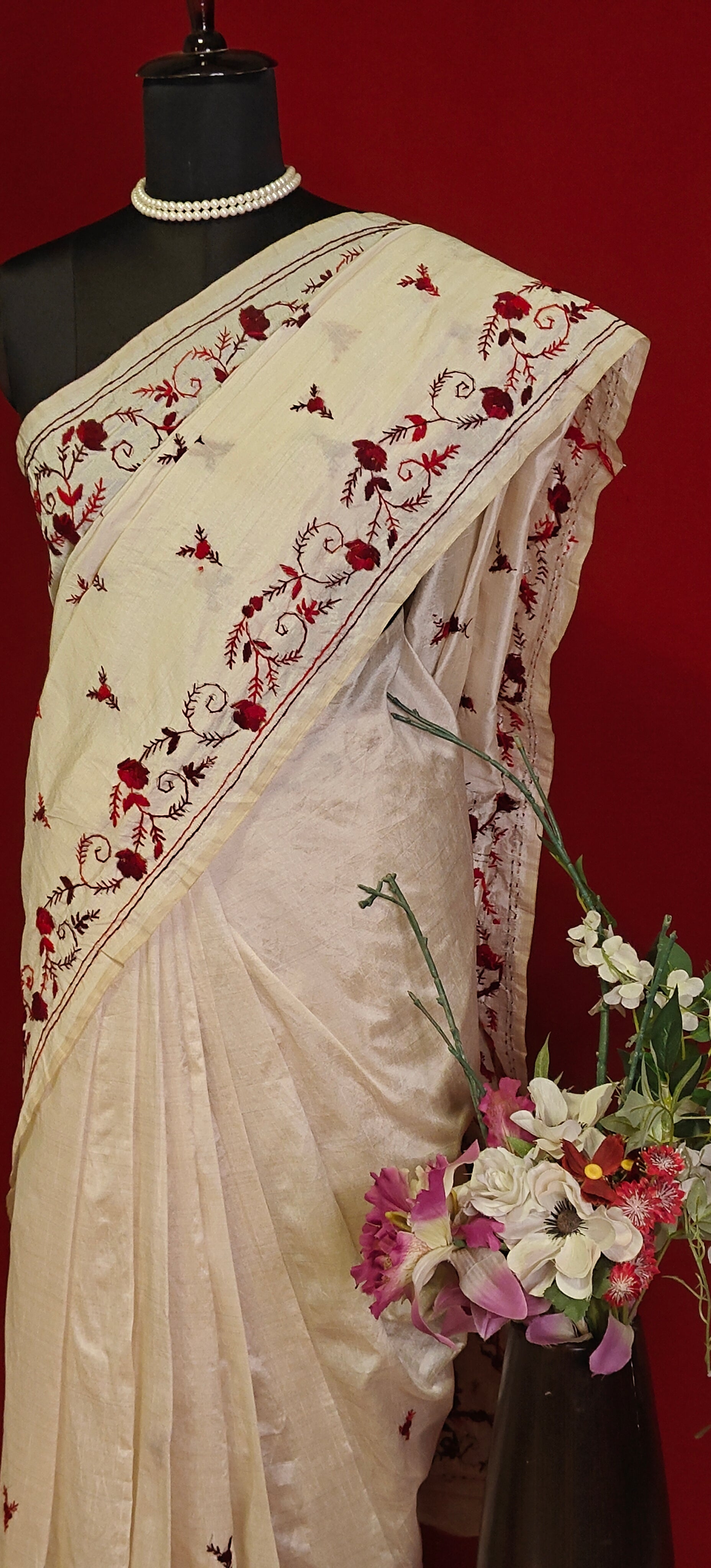 Handcrafted kantha stitched saree in cream with floral embroidery in maroon and red on a mannequin with a flower vase against a red background.