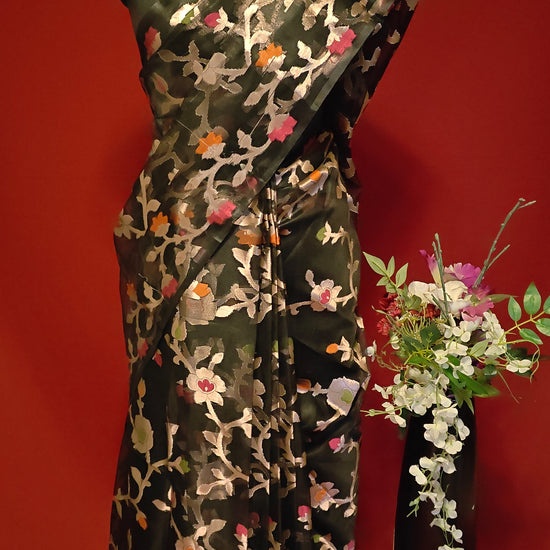 Black muslin blended silk saree with Jamdani weave and gold zari jaal, styled on mannequin with flower vase and red background.