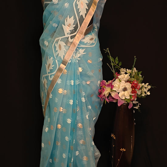 Sky blue organza muslin saree with golden border and Jamdani floral embroidery, styled on mannequin with flower vase against a black background.