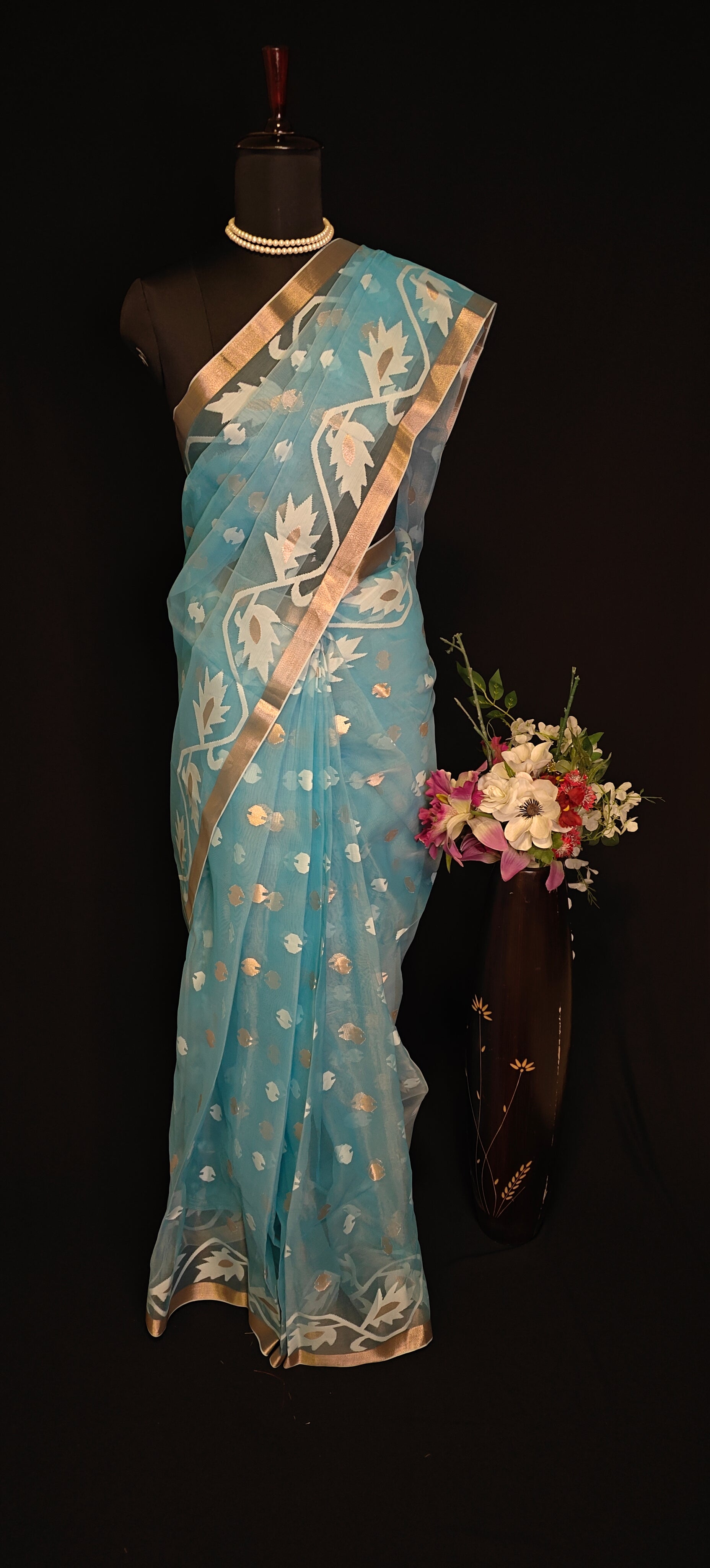 Sky blue organza muslin saree with golden border and Jamdani floral embroidery, styled on mannequin with flower vase against a black background.