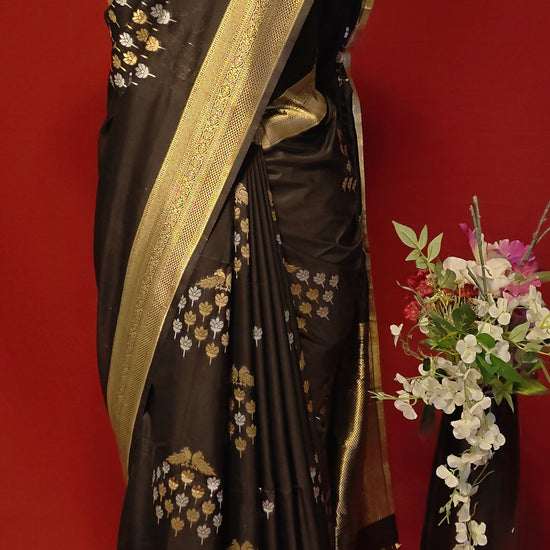 Black pure soft silk saree with gold and silver floral motifs and rich zari border draped on mannequin along with flower vase against a red background.