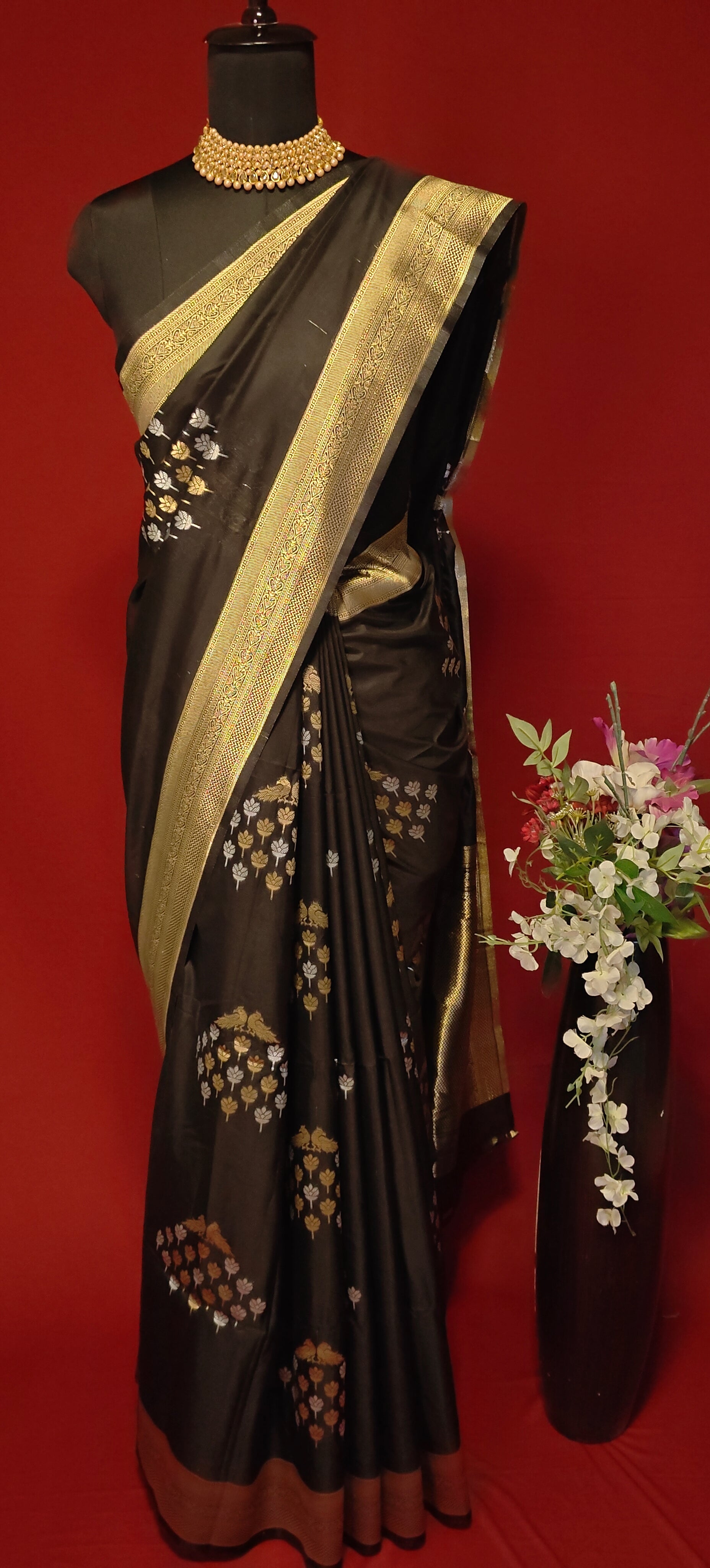 Black pure soft silk saree with gold and silver floral motifs and rich zari border draped on mannequin along with flower vase against a red background.