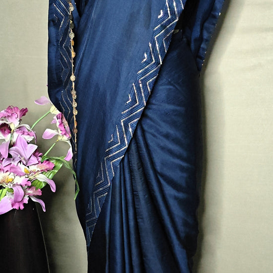 Blue silk saree with metallic zigzag border in gold and silver, displayed on mannequin with pearl necklace.