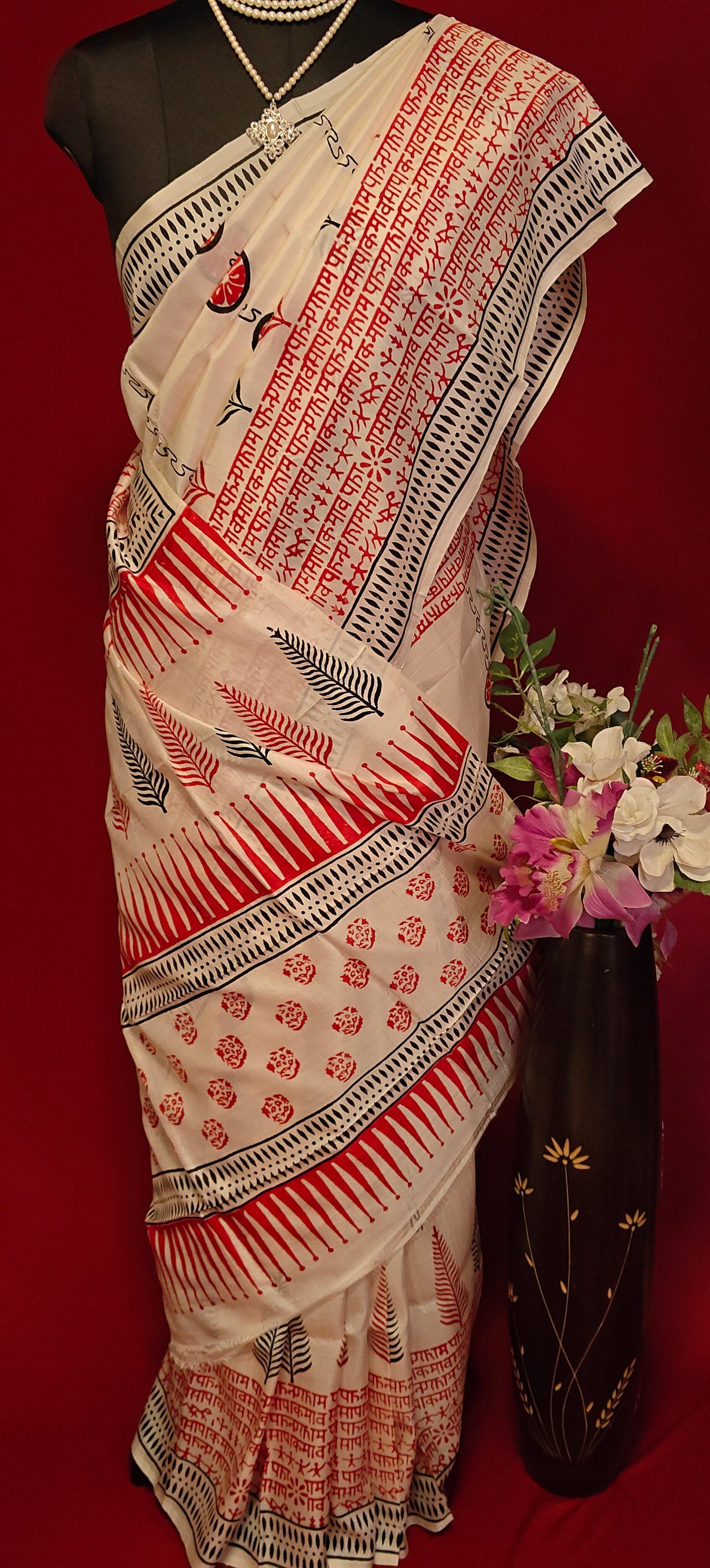 Pure Bishnupuri silk saree in white with red and black printed motifs inspired by traditional Indian art on a mannequin showcasing saree pallu with flower vase against a red background.