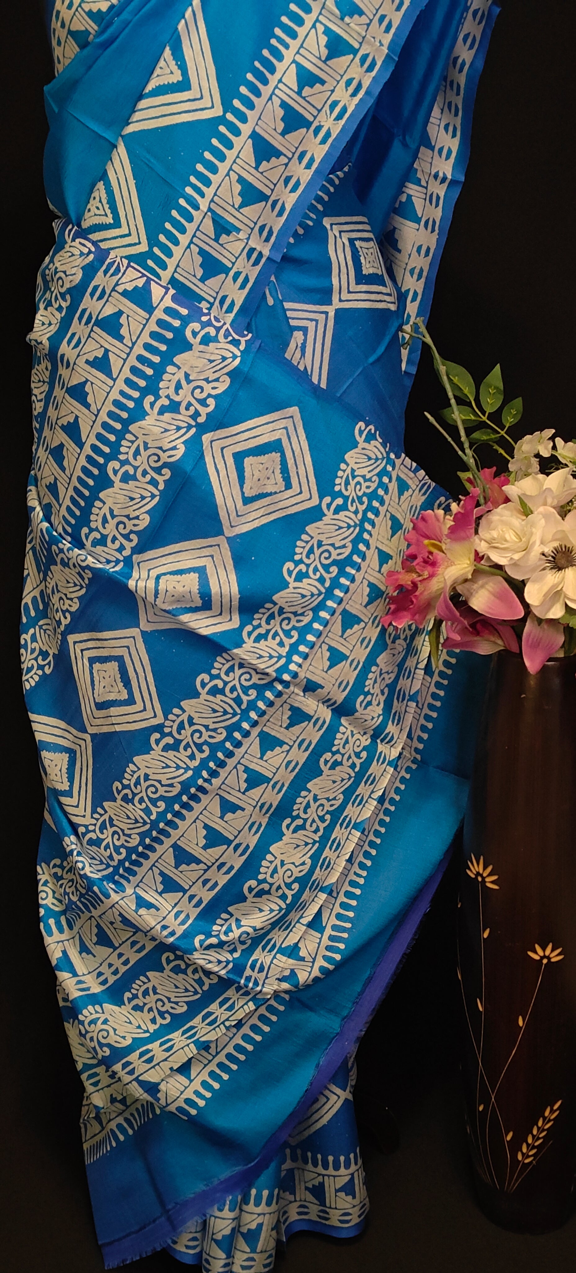 Pure bishnupuri silk saree in royal blue with white digital print motifs and geometric border, styled on mannequin with flower vase, showcasing the pallu of the saree against black background.