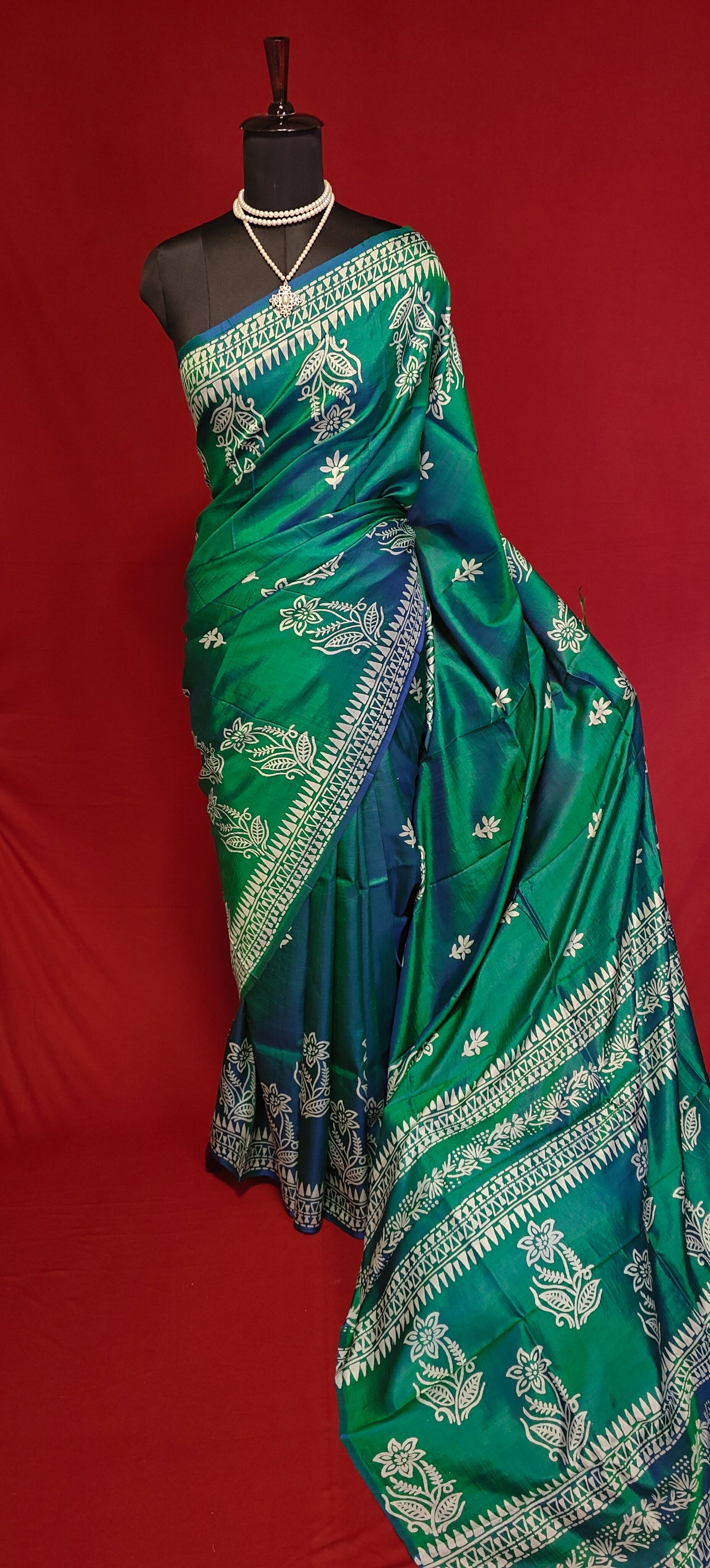 Pure bishnupuri silk saree in dual tone green and blue with white floral motifs and elegant border draped on a mannequin against red background.