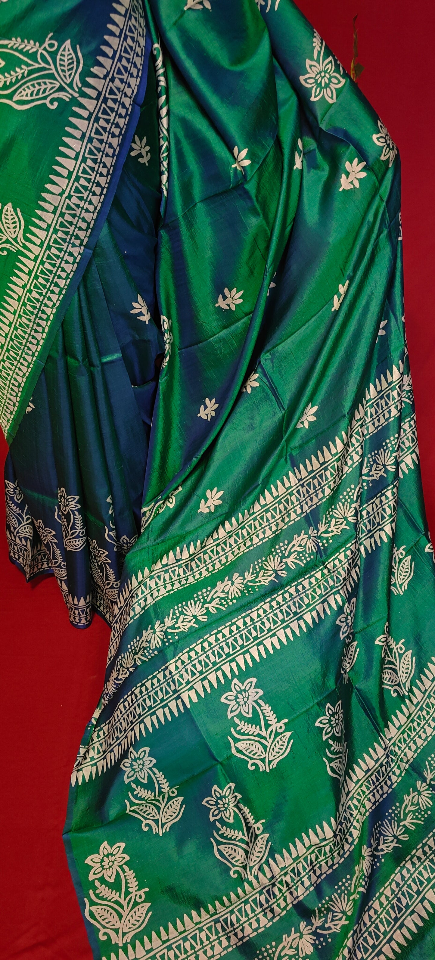 Pure bishnupuri silk saree in dual tone green and blue with white floral motifs and elegant border draped on a mannequin showcasing the pallu against red background.