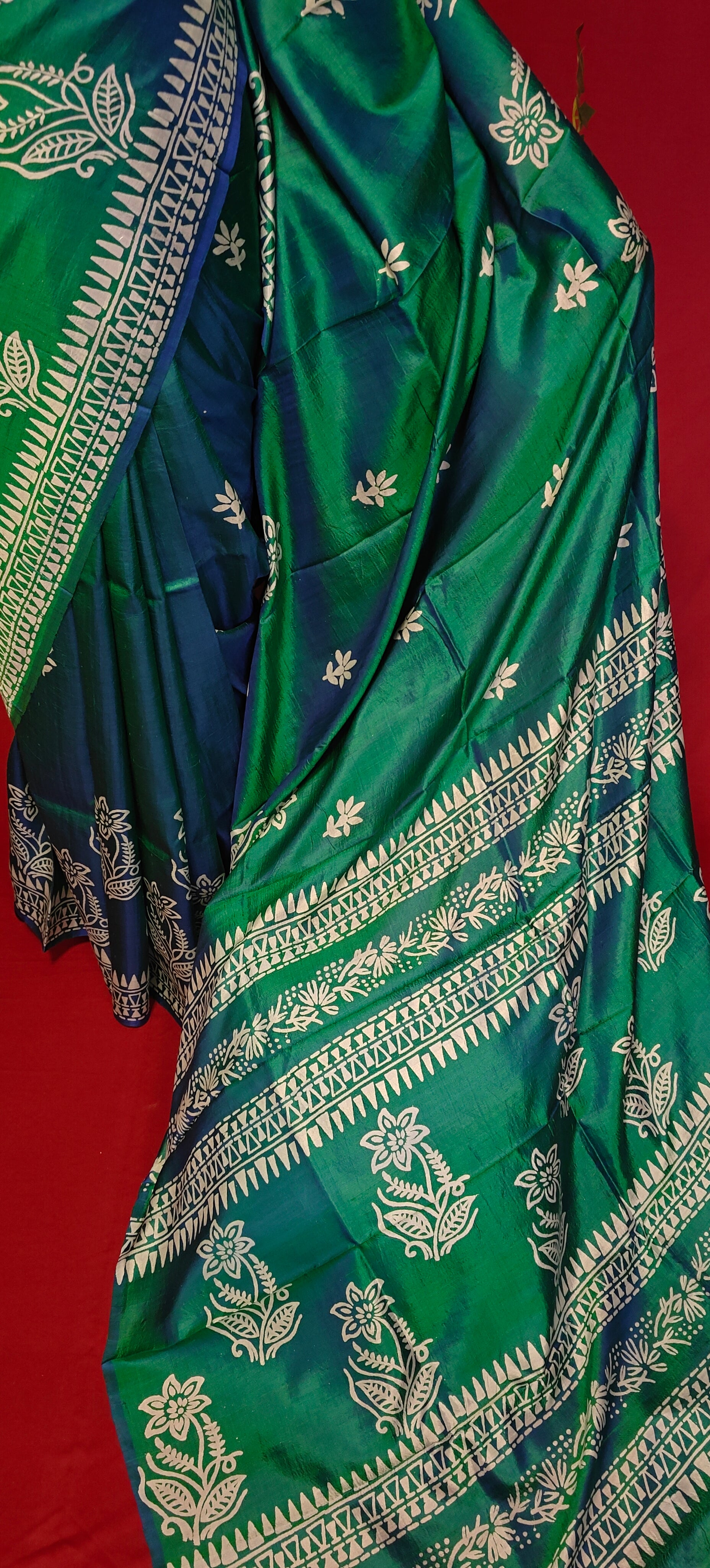 Pure bishnupuri silk saree in dual tone green and blue with white floral motifs and elegant border draped on a mannequin showcasing the pallu against red background.