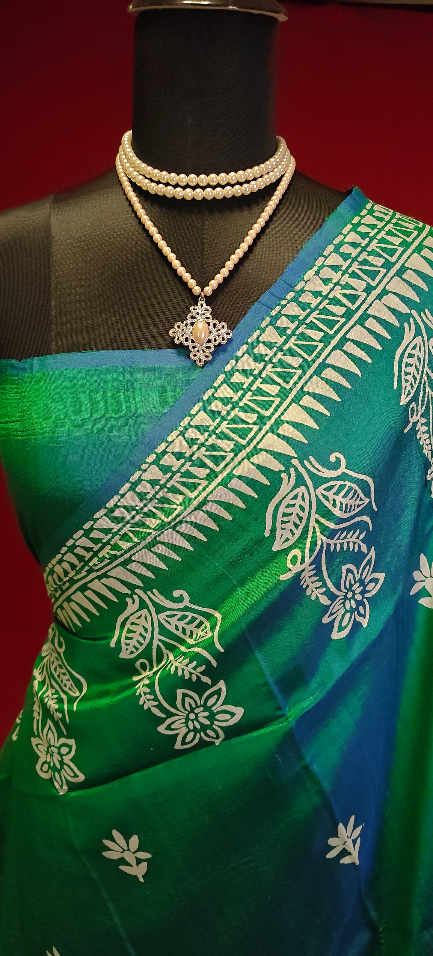 Pure bishnupuri silk saree in dual tone green and blue with white floral motifs and elegant border draped on a mannequin showcasing the blouse piece against red background.