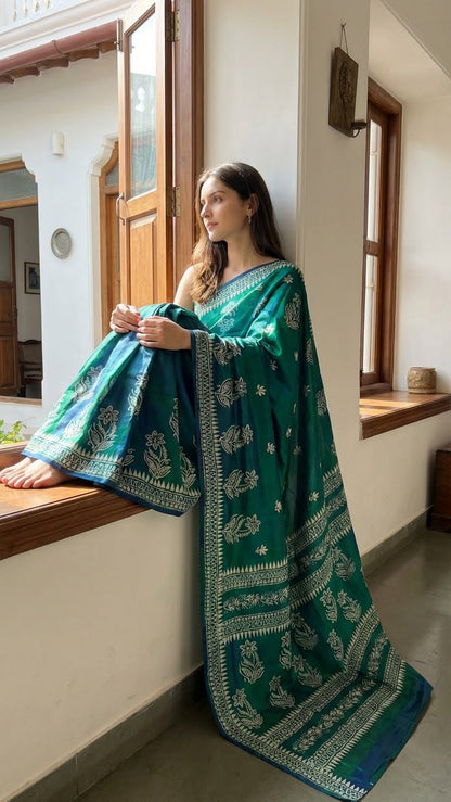 Woman wearing a pure Bishnupuri silk saree in dual tone green and blue with white floral motifs and elegant border draped with a traditional design, sitting by a window.