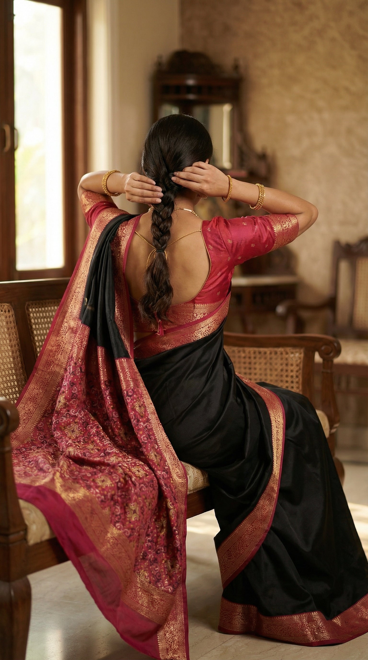 Woman wearing black pure soft silk saree with magenta zari border and golden motifs, sitting with her back to the camera, hands raised to braid her hair, which fully reveals the back-neck design of the blouse.