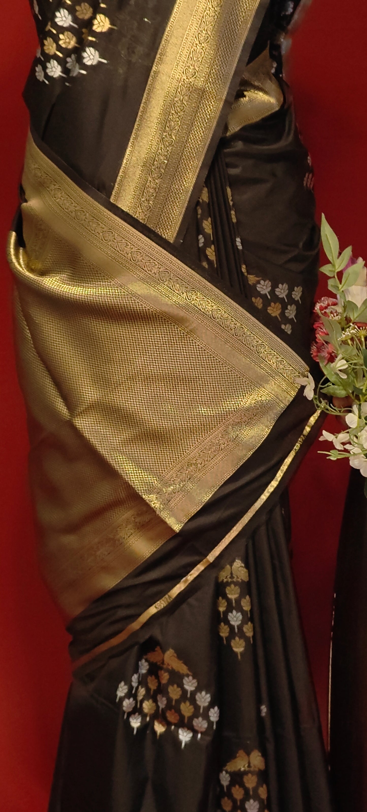 Black pure soft silk saree with gold and silver floral motifs and rich zari border draped on mannequin showcasing the rich pallu with zari work along with flower vase against a red background.