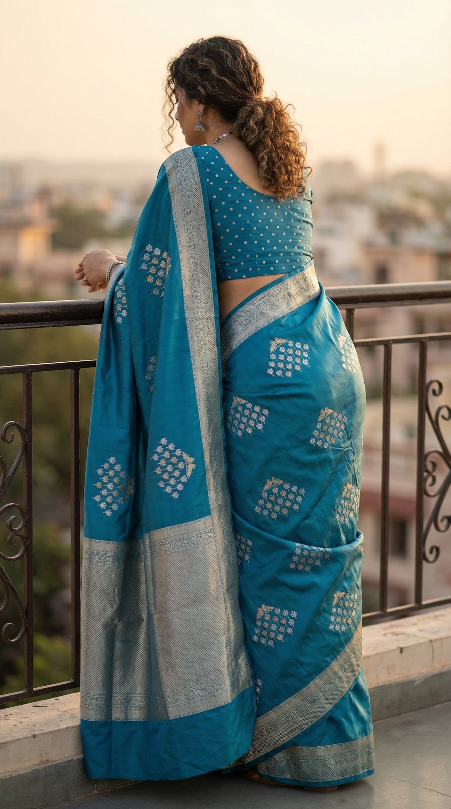 Woman wearing Blue pure soft silk saree with silver leaf motifs and luminous zari detailing. Leaning forward on a railing with the back to the camera; the pallu hangs vertically, showing the full length of the border design.