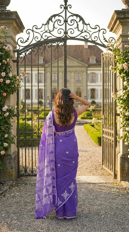 Woman in a purple-applique-kota-saree, standing in front of an ornate iron gate with a grand building in the background.
