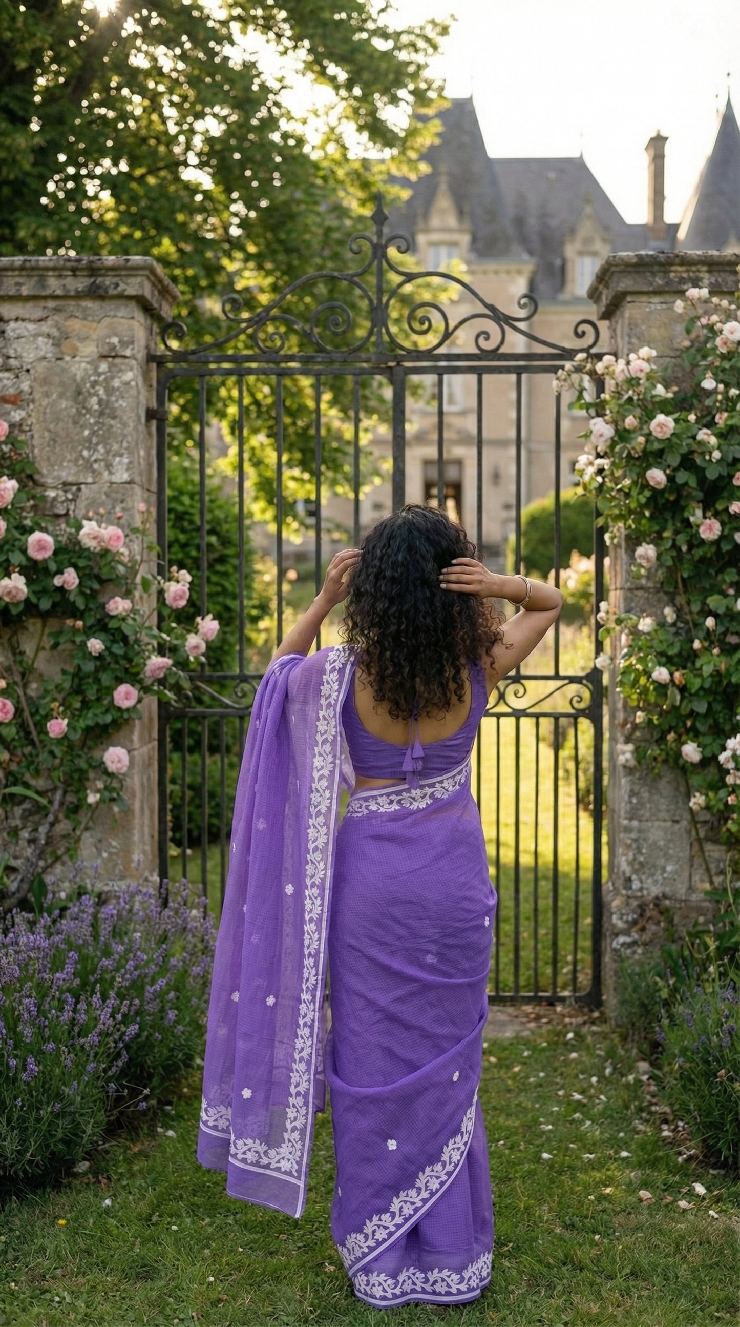Woman in a purple-kota-doria-saree, standing in front of a chateau with floral gates.