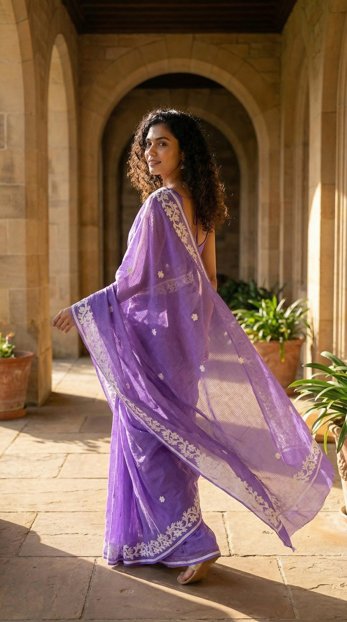 Woman in a purple-kota-doria-saree, standing in a sunlit outdoor setting.