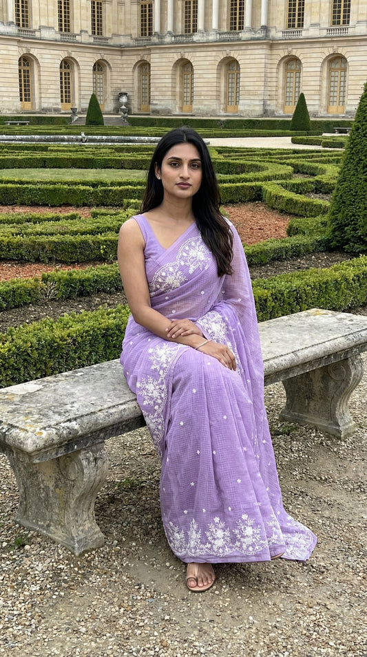 Woman in a purple-resham-kota-cotton-saree, sitting on a stone bench in a garden.