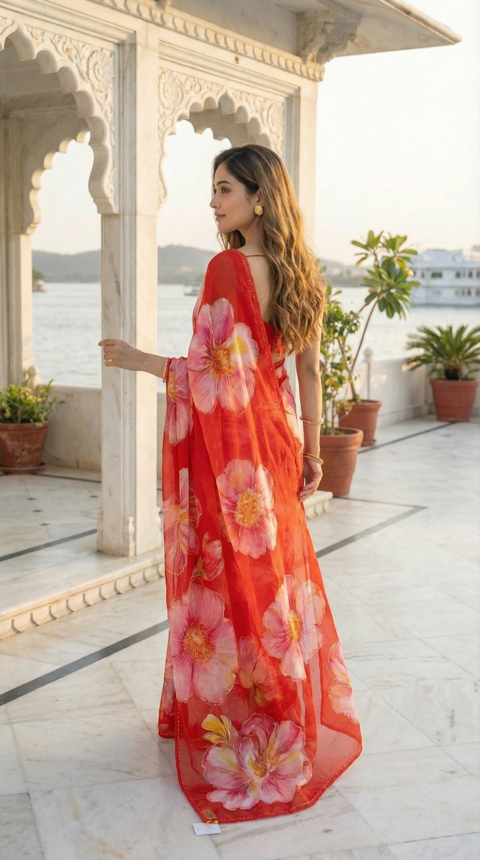 Woman in a red chiffon saree with large floral prints, standing on a balcony with plants and a scenic view.