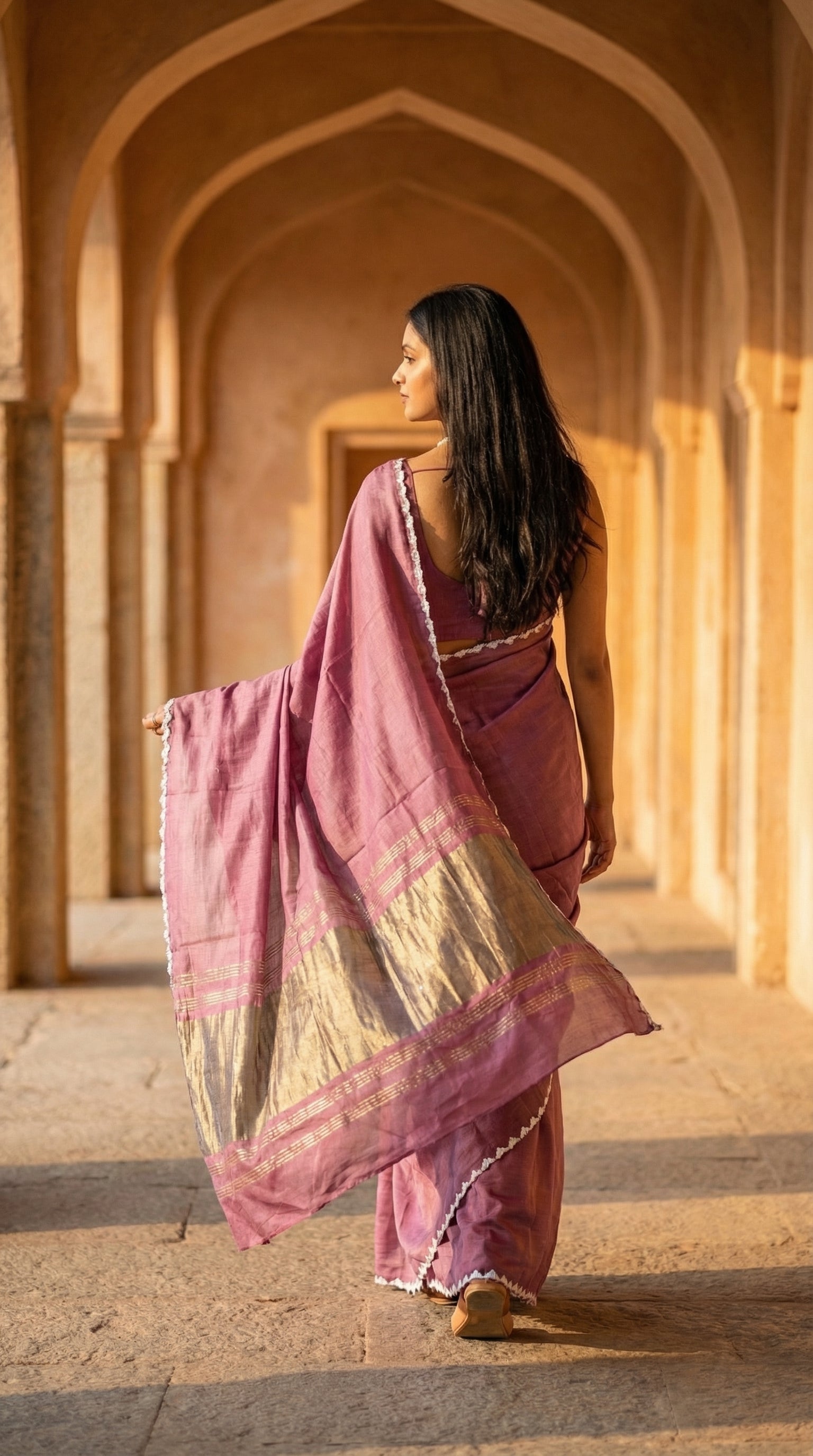 Woman in a rose-pink-modal-silk-saree-lagdi-patta-pallu, back-facing and walking down the corridor of an architectural setup.