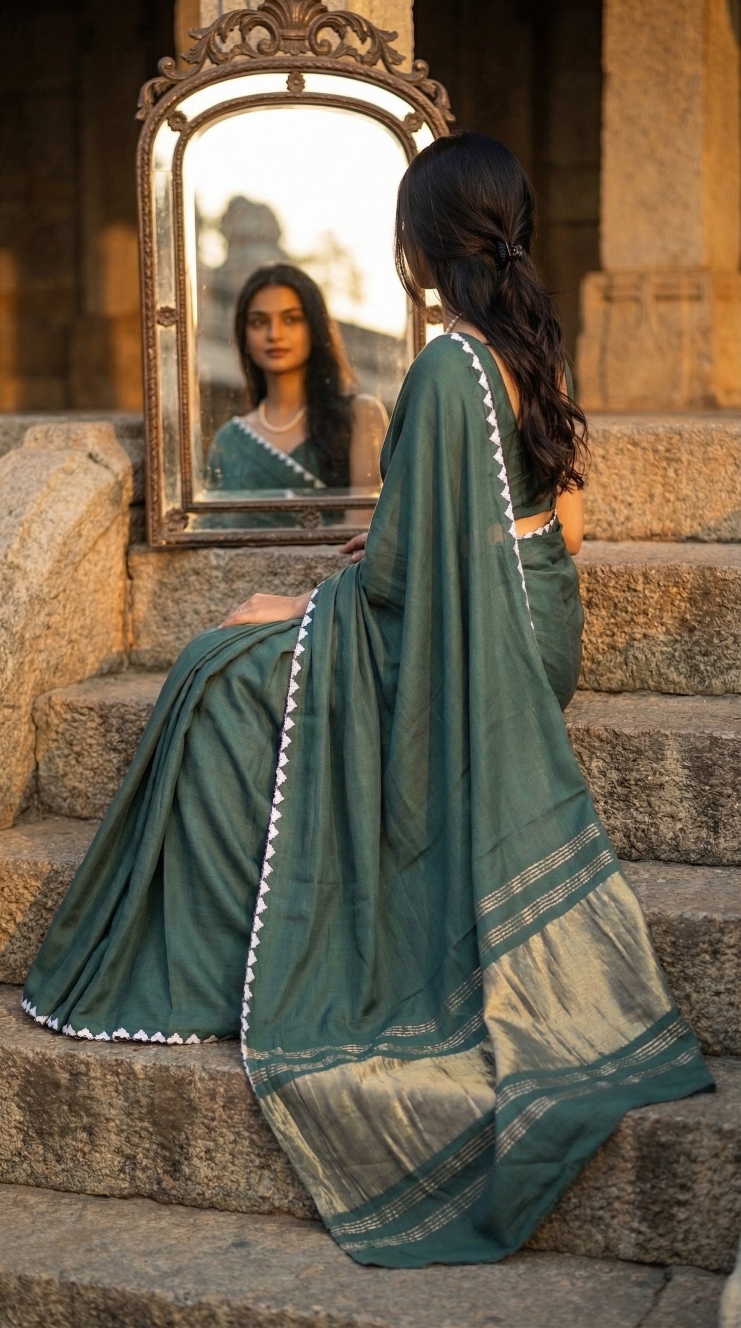 Woman in a sage-green-modal-silk-saree-lagdi-patta-pallu, sitting on steps, looking at herself in a mirror.