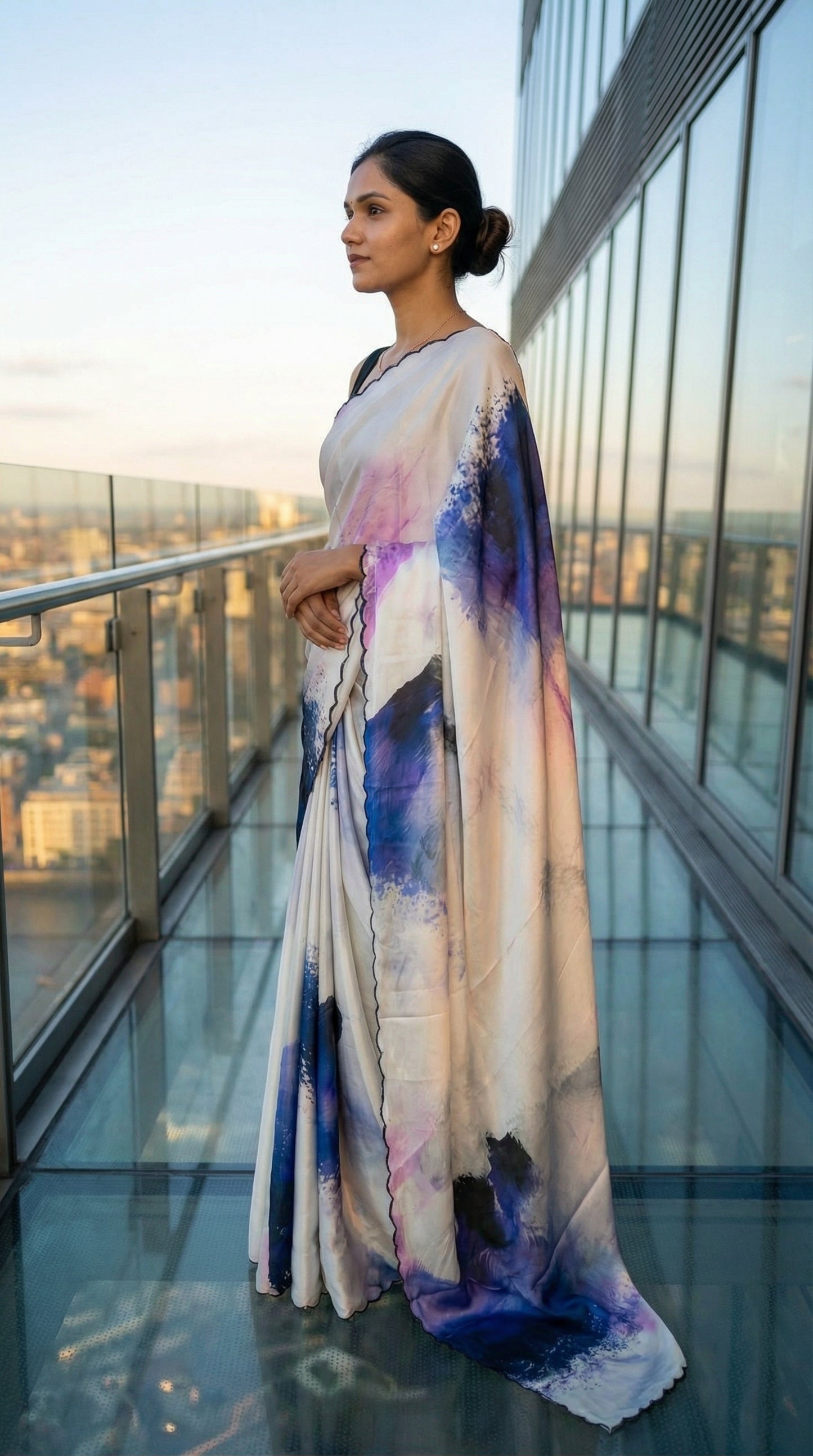 White satin-georgette saree with blue abstract brushstroke design and scalloped embellished border, on a rooftop with cityscape view.