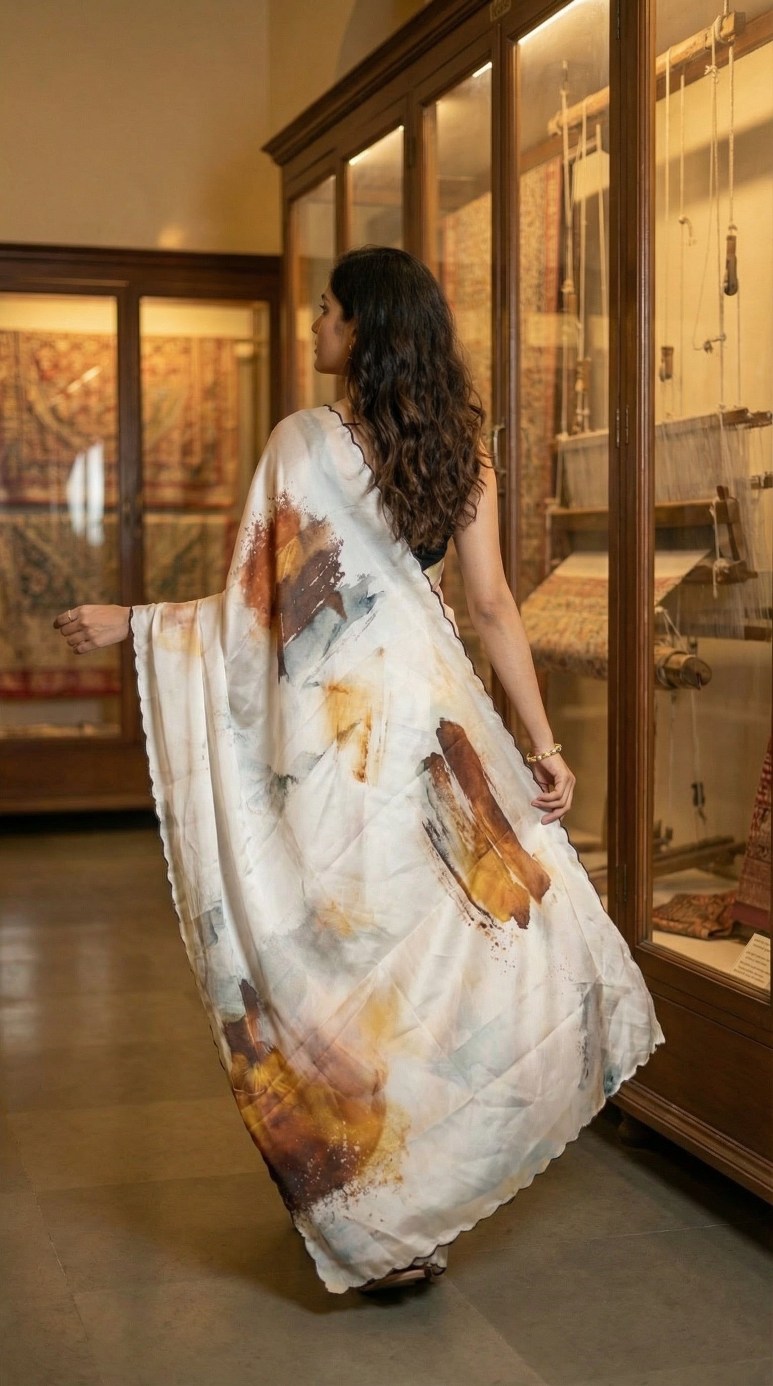 Woman in a brown-accented satin-georgette saree featuring artistic brushstroke patterns on white base with scalloped edge, in an indoor setting.