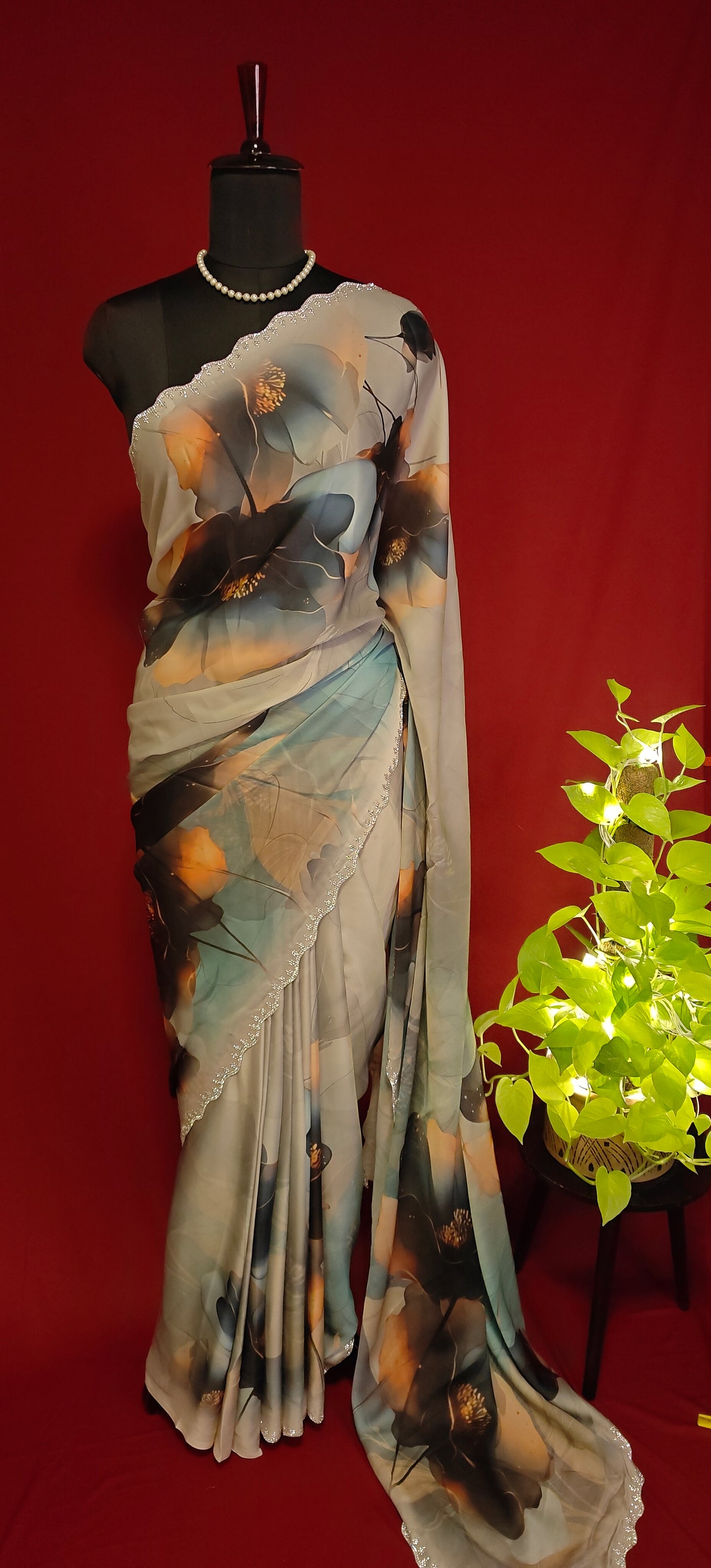 Cream satin georgette saree with oversized floral print and silver scalloped border, styled on mannequin against a red background with a plant to the side.