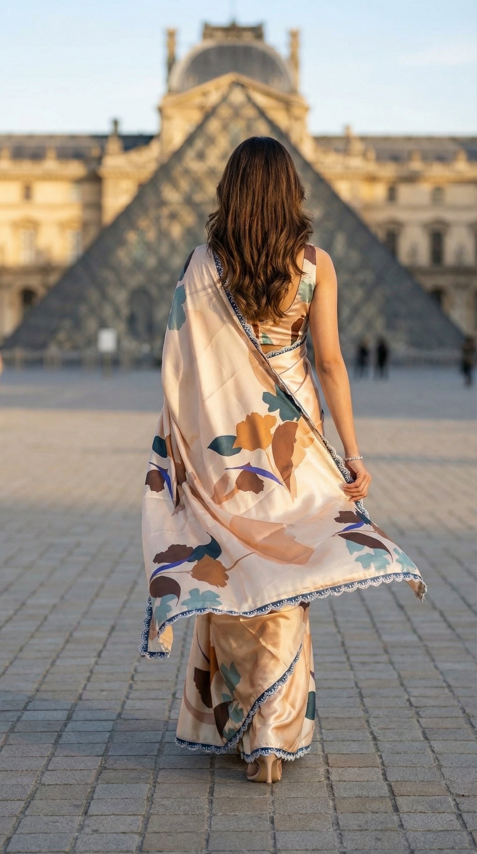 Woman wearing a Peach satin-georgette saree featuring bold floral motifs and delicate lace trim, in front of the Louvre Pyramid in Paris.