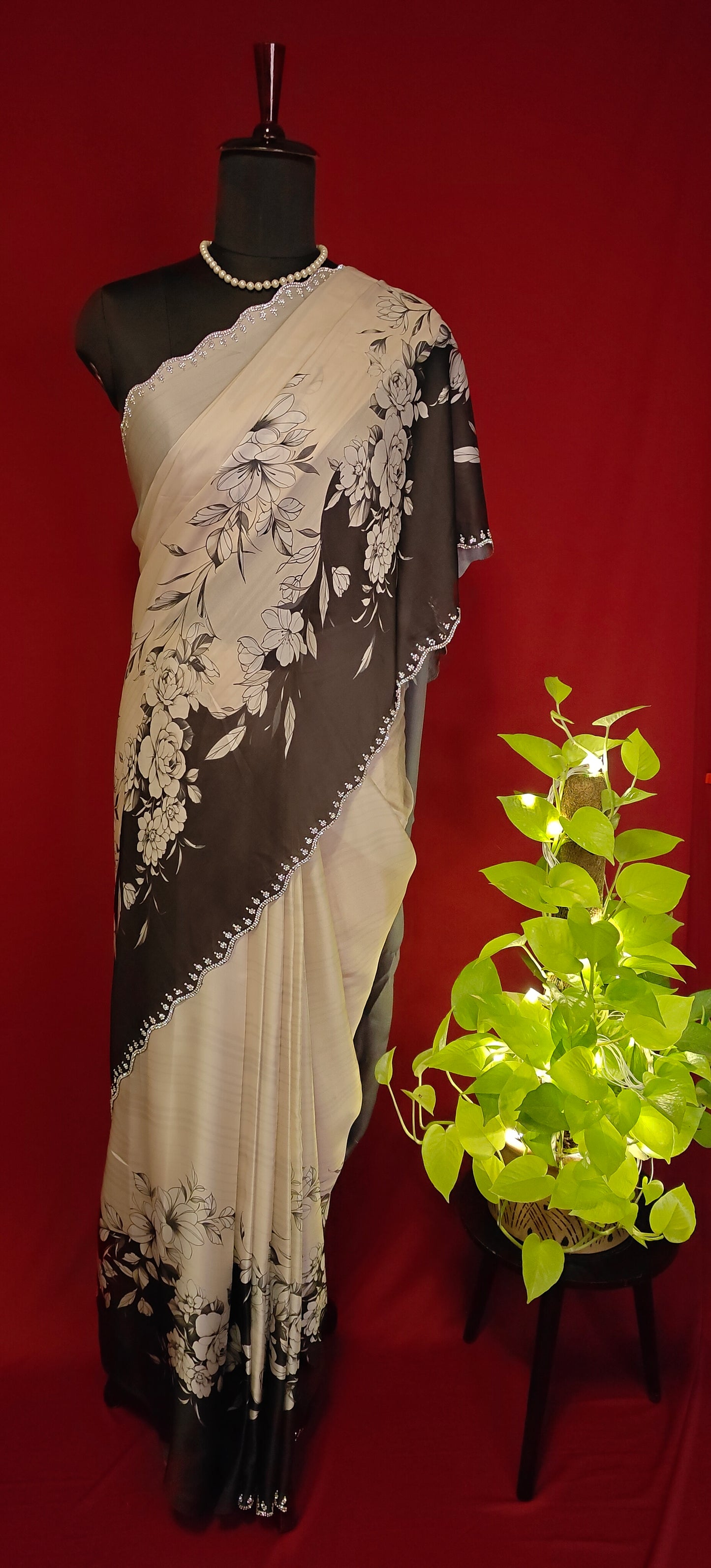 Black and white satin saree with abstract floral print and white pallu, styled on mannequin with a flower vase illuminated with lights on a table against red background.