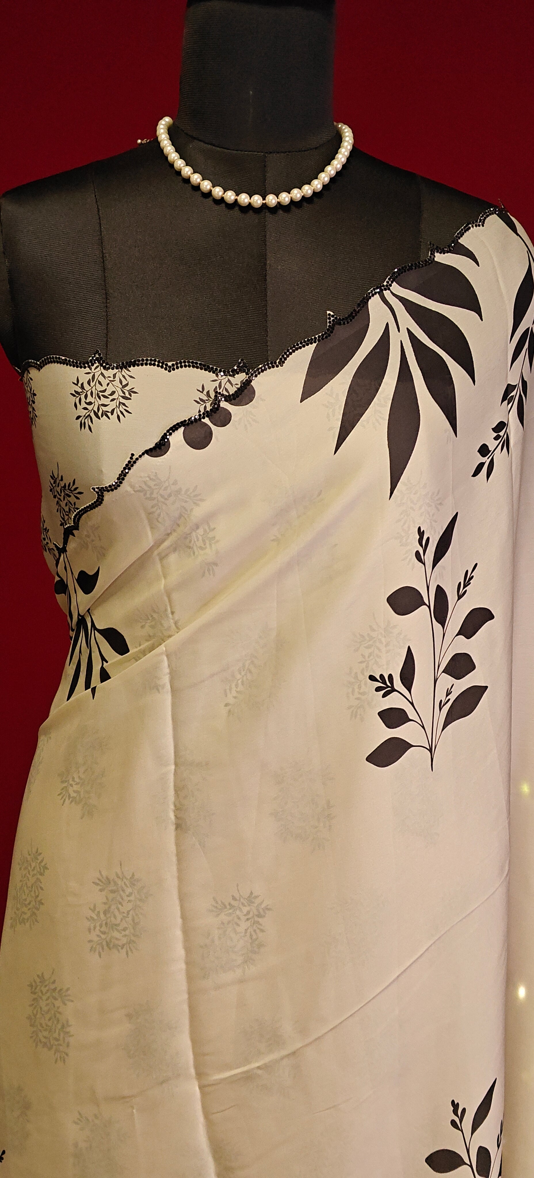 Cream satin saree with black leaf print and black motif border, styled on mannequin showcasing the blouse piece against a red background.