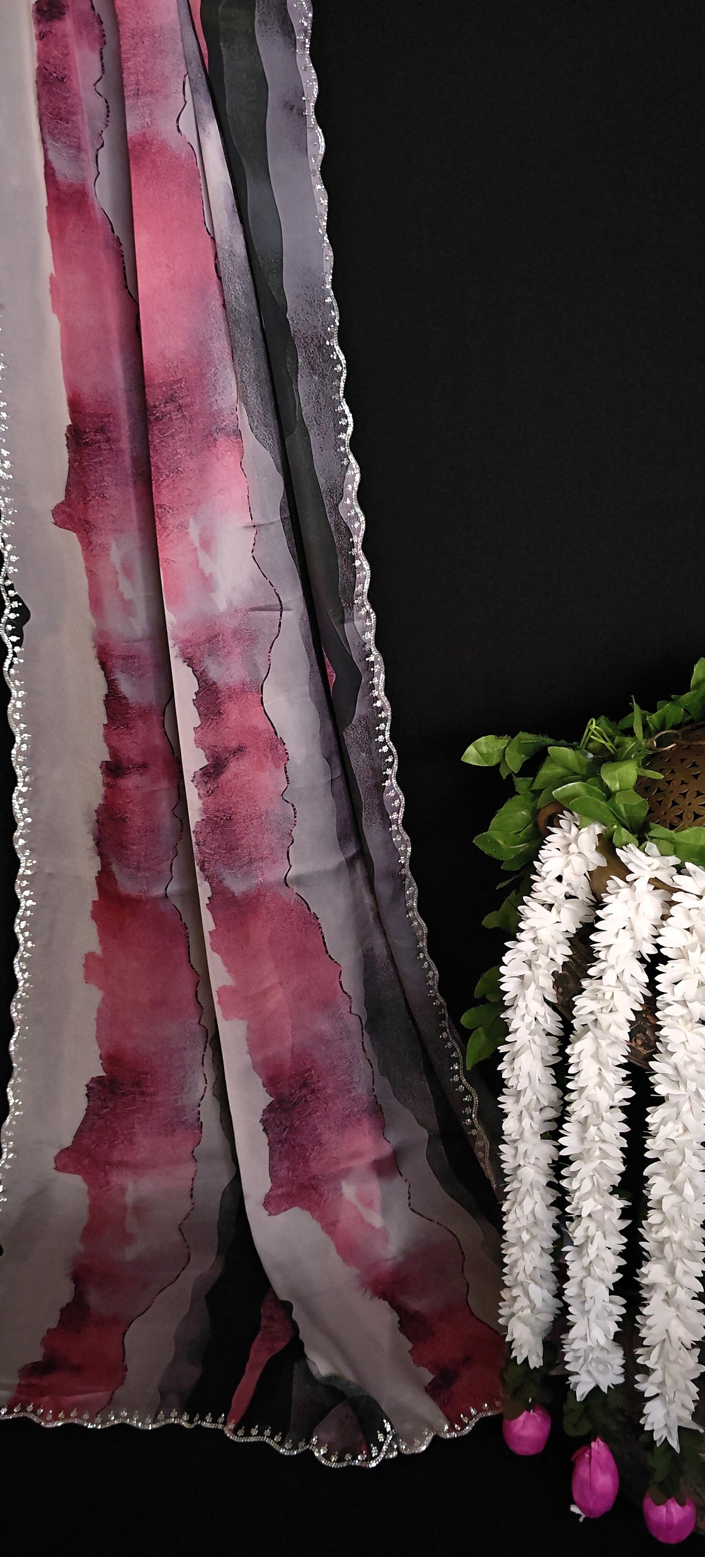 Pink satin saree with abstract print and scalloped silver border next to a plant with white flowers on a black background.