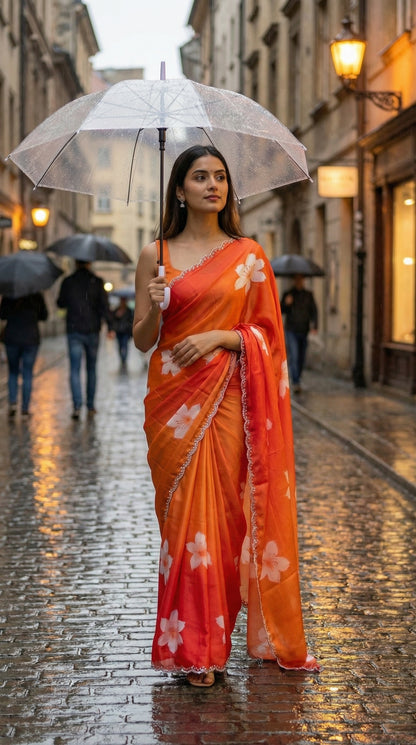 Woman wearing a red to orange gradient satin saree with white floral motifs and light Zari border, holding a transparent umbrella on a rainy street.