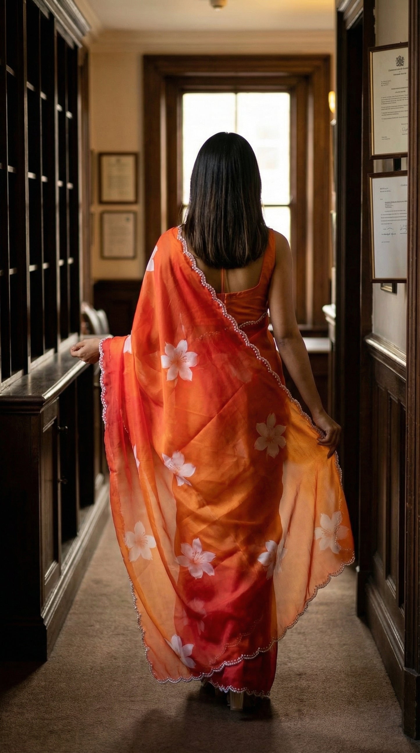 Woman wearing a red to orange gradient satin saree with white floral motifs and light Zari border, walking through a hallway with wooden paneling.