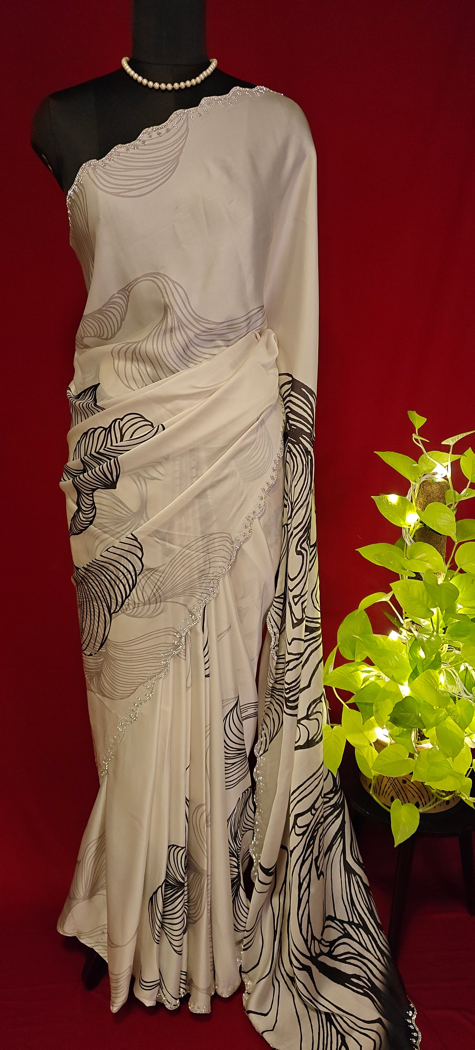 White satin saree with black abstract design and silver motif border, styled on mannequin with plant put on a table against red background.