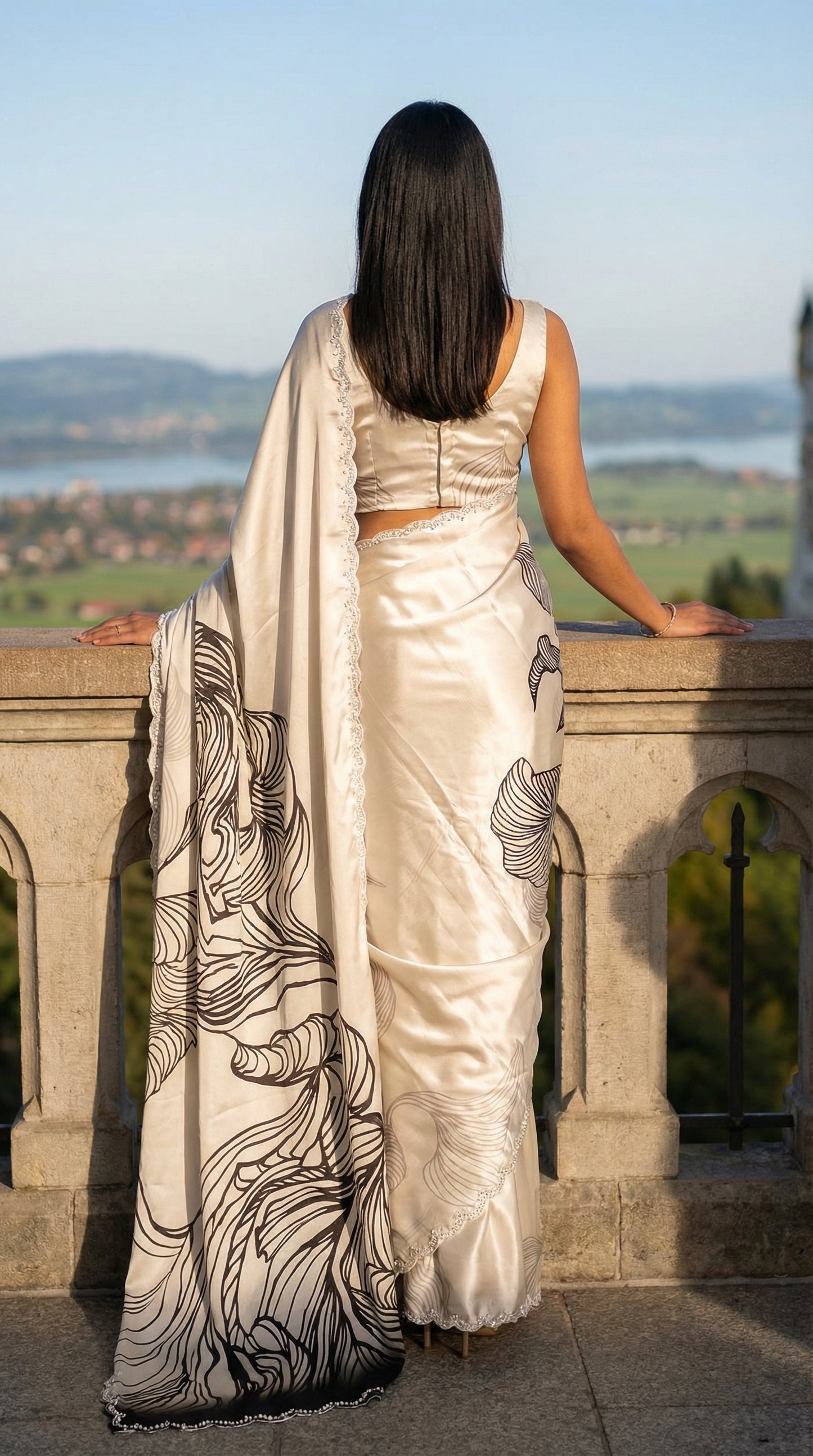 Woman in a White satin saree with black abstract design and silver motif border, standing on a balcony overlooking a scenic landscape.