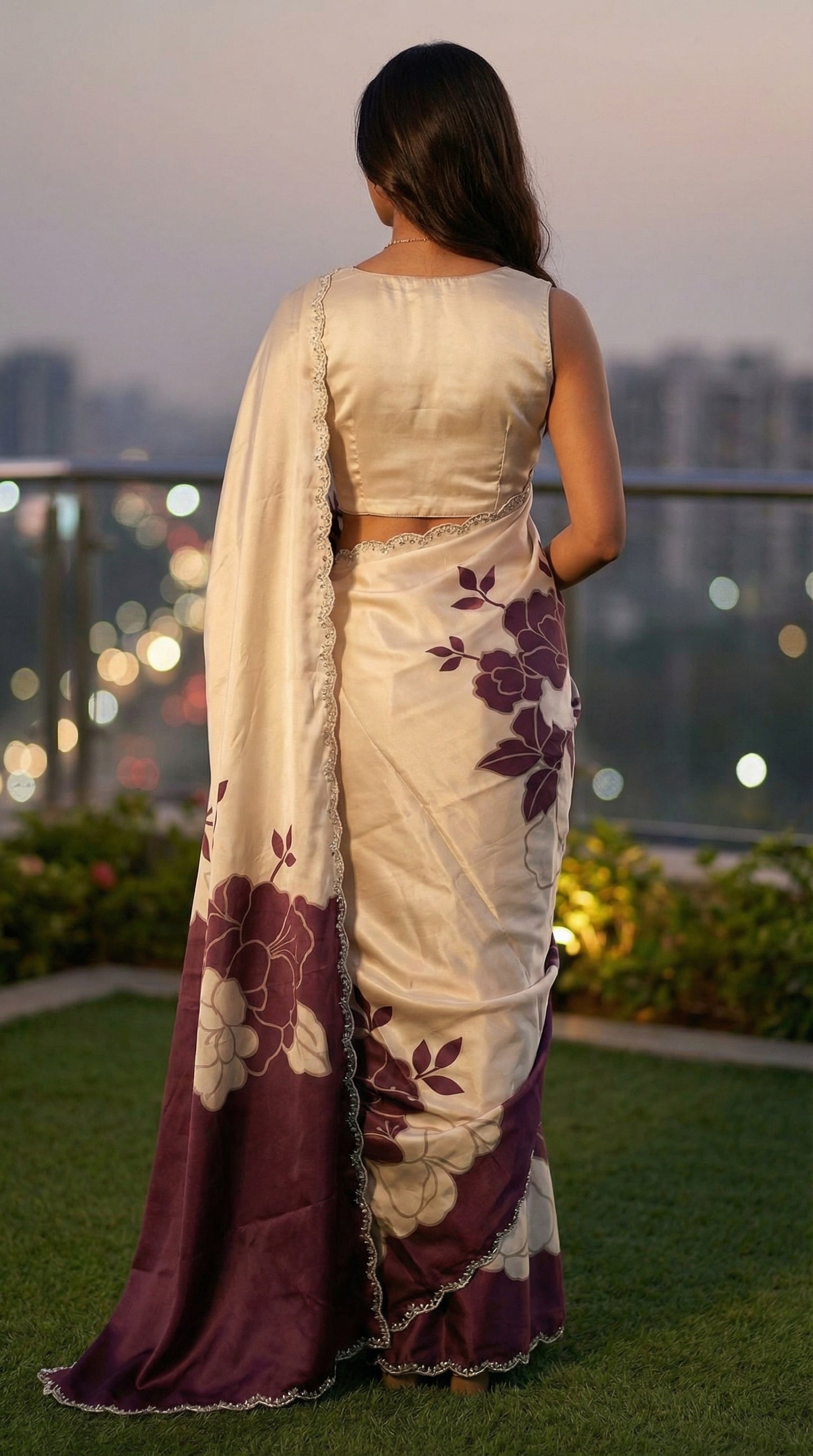 Woman in a Wine red satin saree with large floral patterns and scalloped border, standing on a rooftop with cityscape in the background.