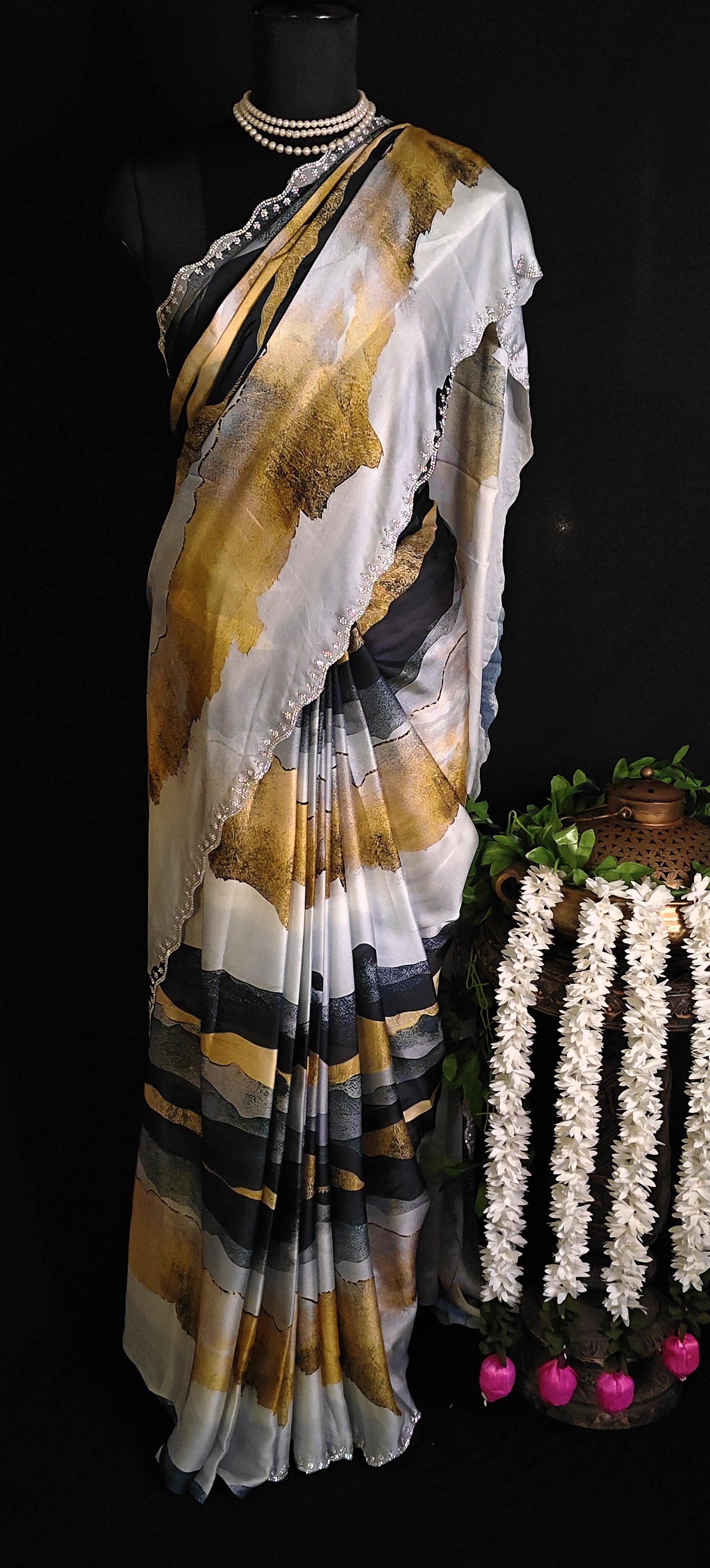 Yellow satin saree with abstract print and scalloped silver border on a mannequin with flowers and a showpiece against black background.