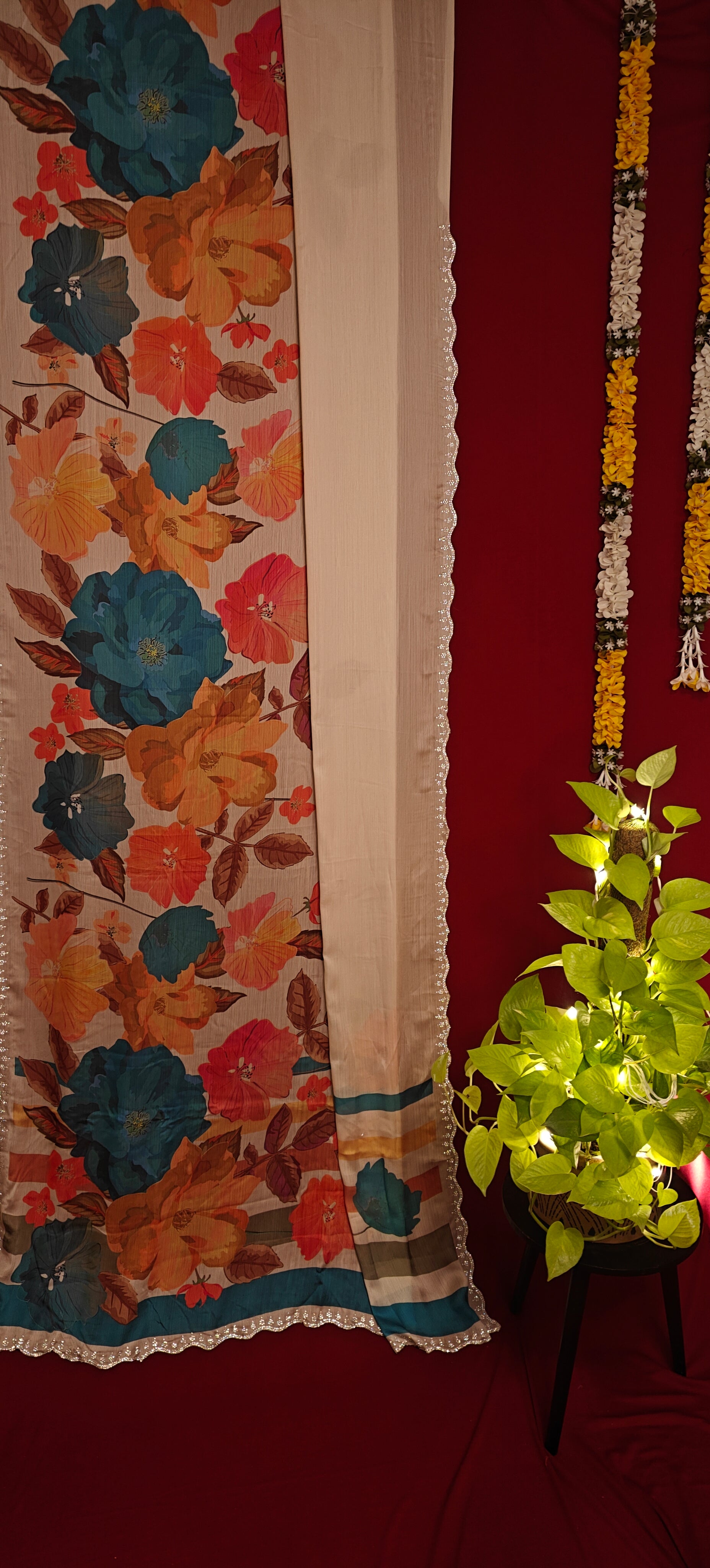 Cream satin chiffon saree with blue floral print and silver scalloped border hanging along with decorative items against red background.