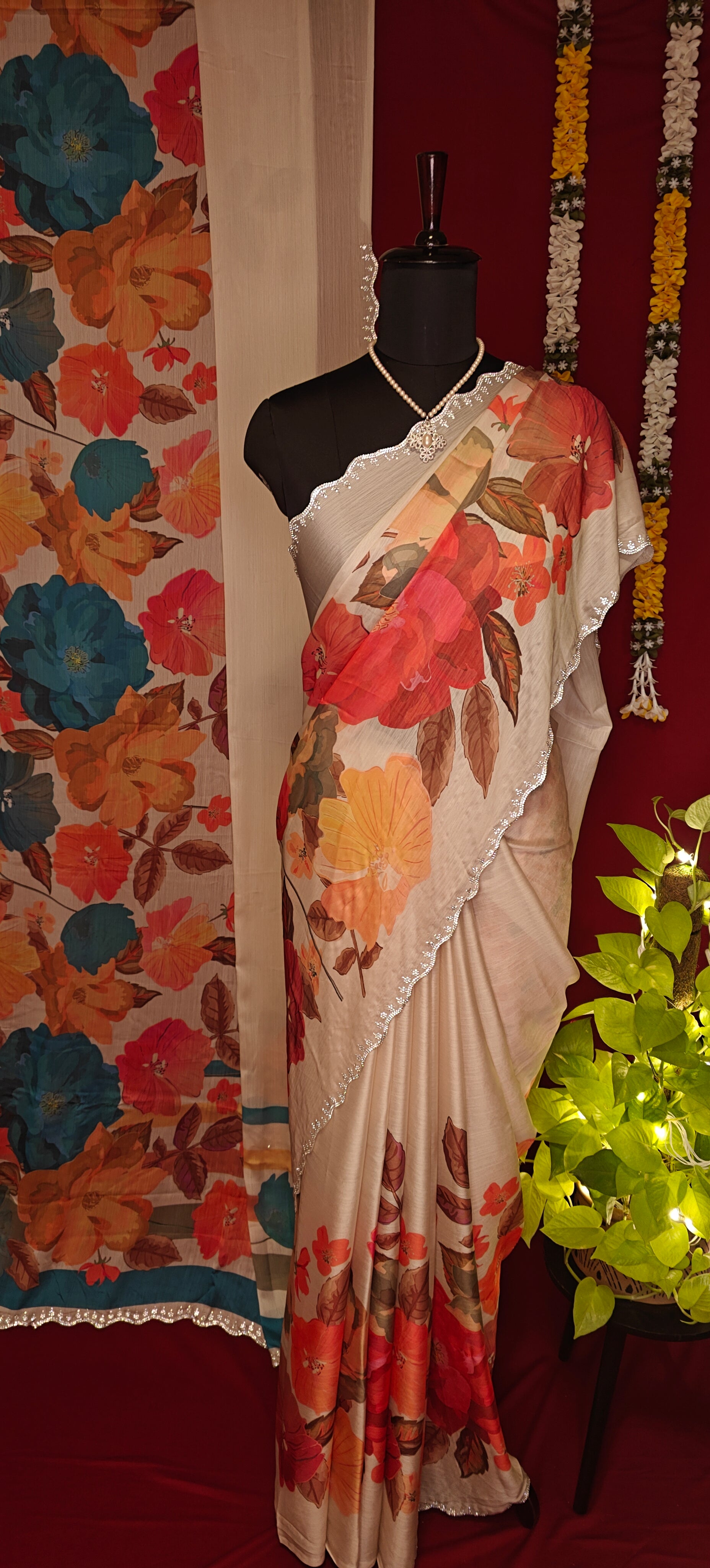 Cream satin chiffon saree with blue floral print and silver scalloped border hanging along with a similar saree with orange floral print put on a mannequin along with decorative items against red background.