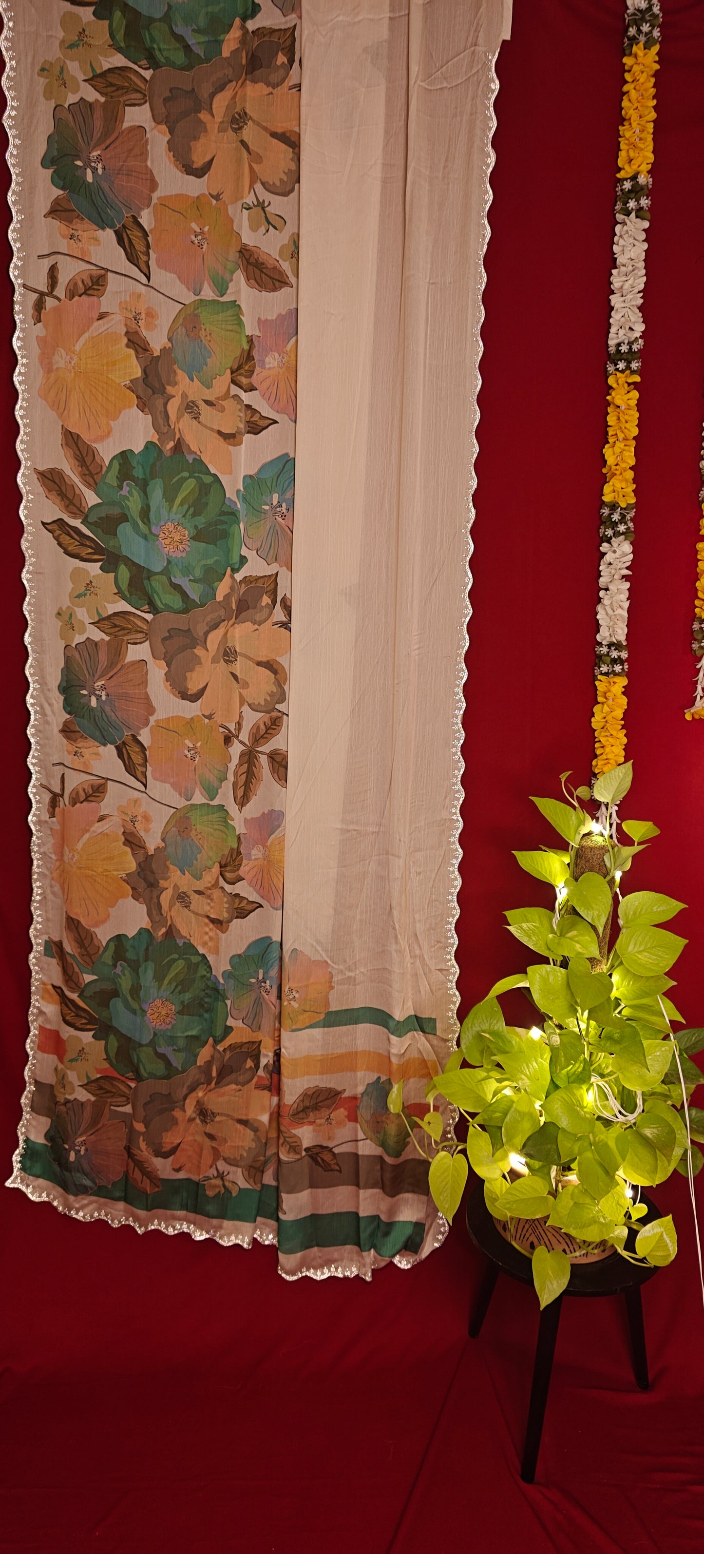 Cream satin chiffon saree with green floral print and silver scalloped border hanging next to a plant against red background.