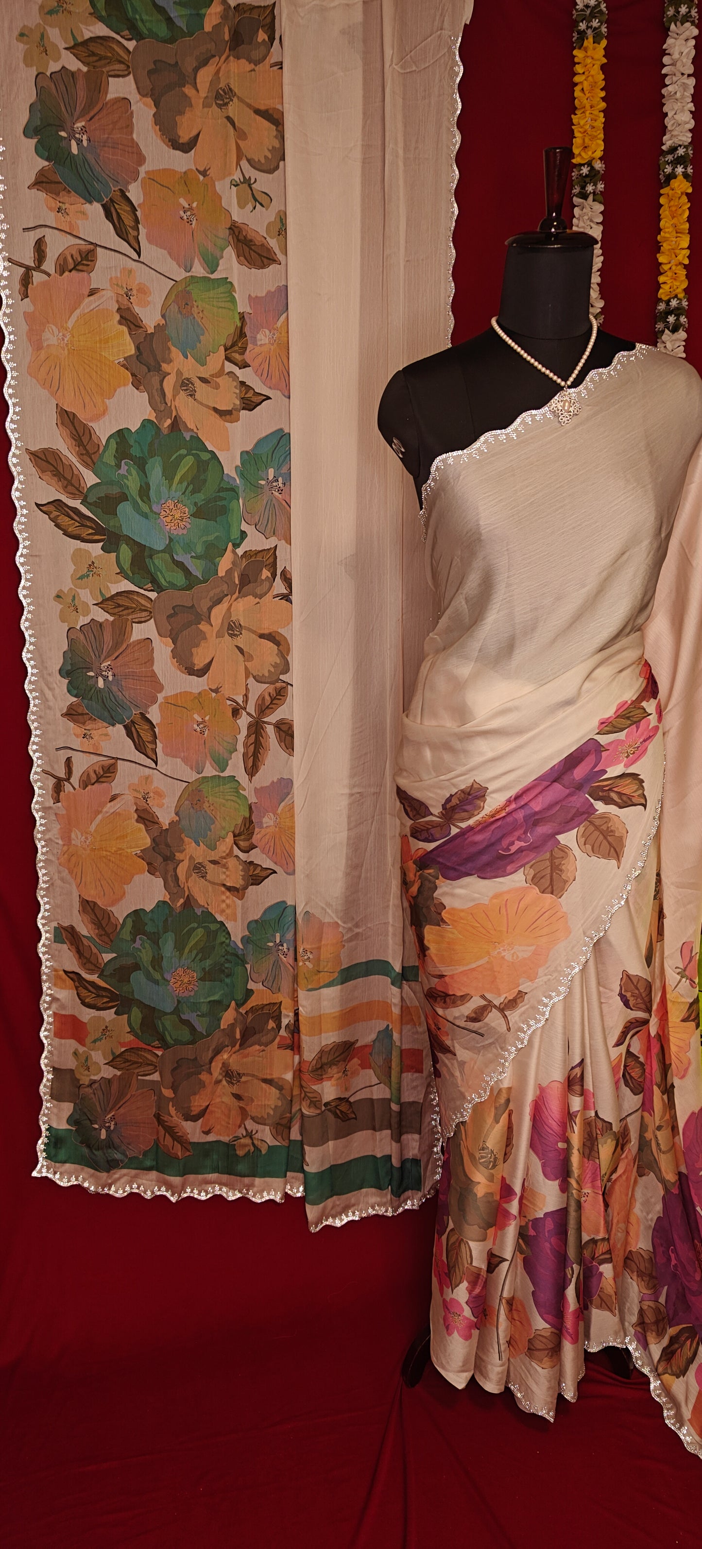 Cream satin chiffon saree with green floral print and silver scalloped border hanging next to a similar saree with purple floral print staged on a mannequin against red background.