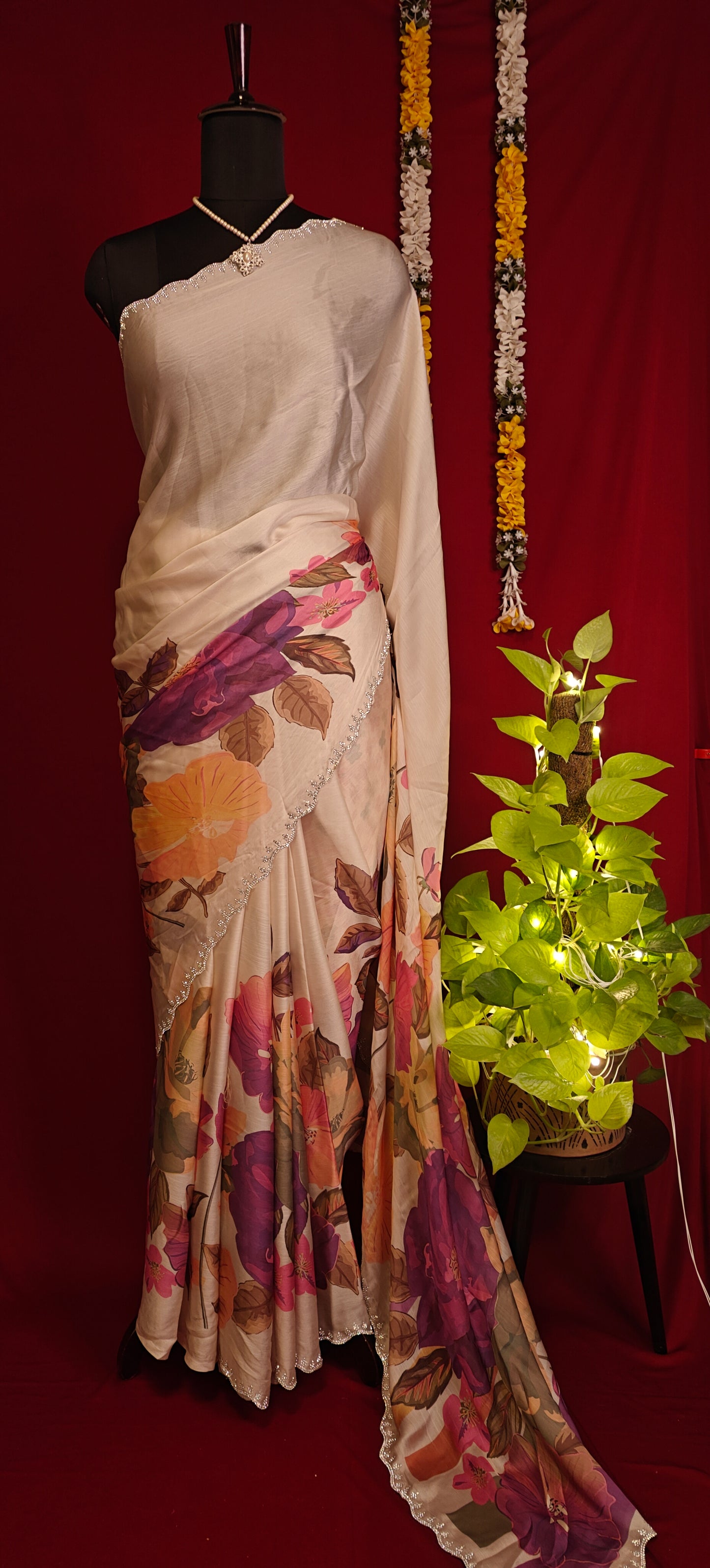 Cream satin chiffon saree with purple floral print and silver scalloped border staged on a mannequin with a plant against red background.
