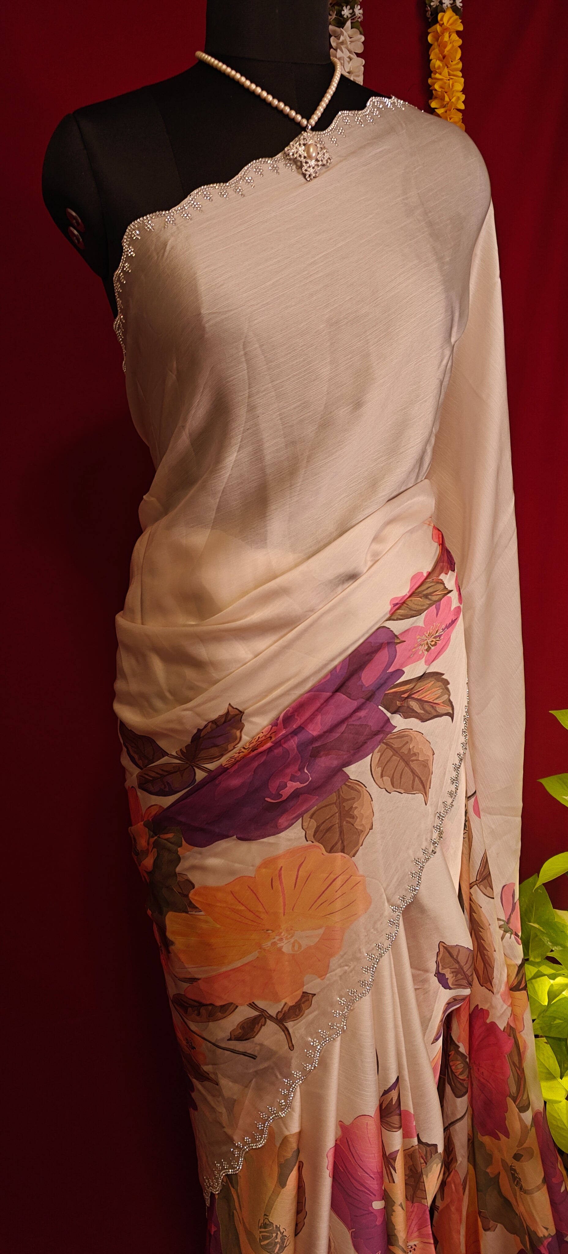 Cream satin chiffon saree with purple floral print and silver scalloped border staged on a mannequin with a plant against red background.