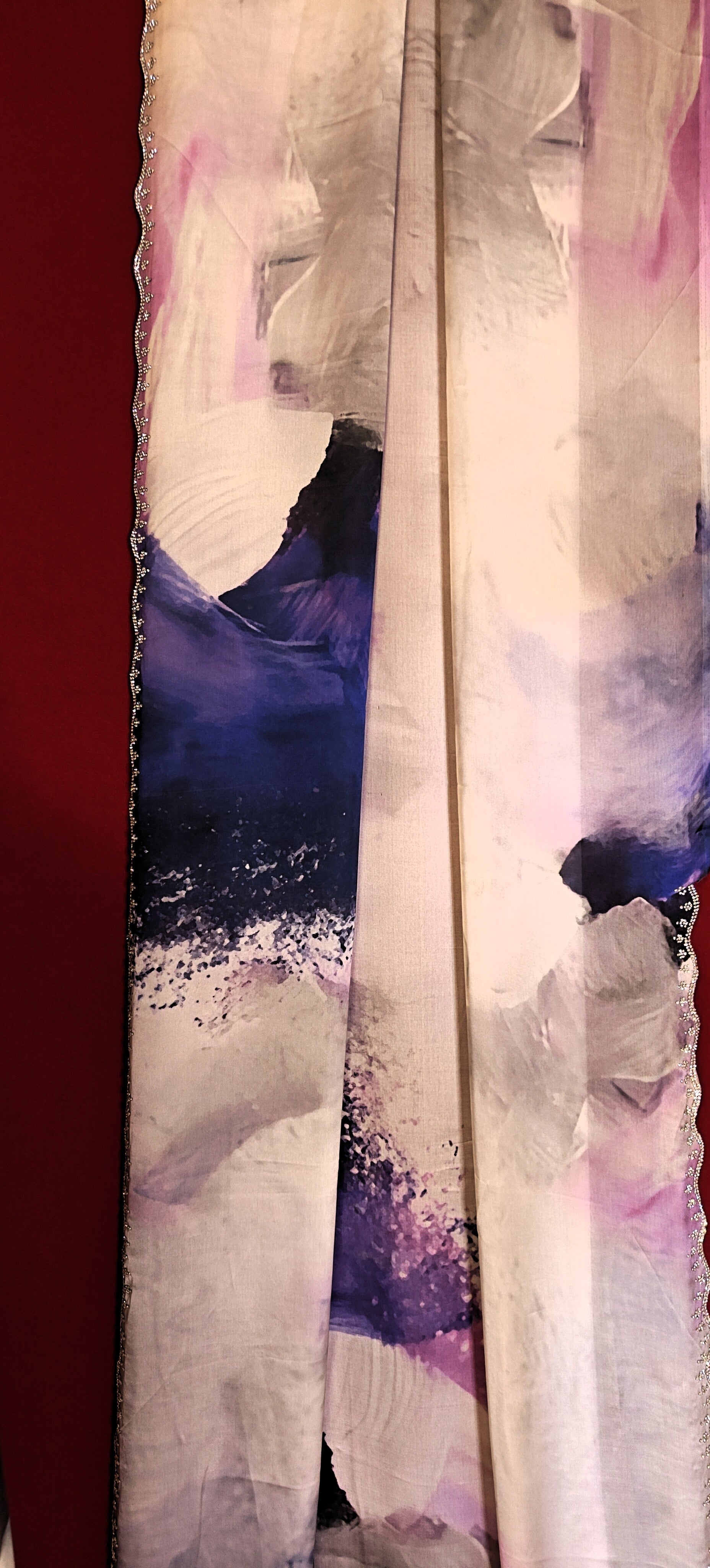 White satin georgette saree with abstract blue brushstroke print and scalloped border hanged against red background.