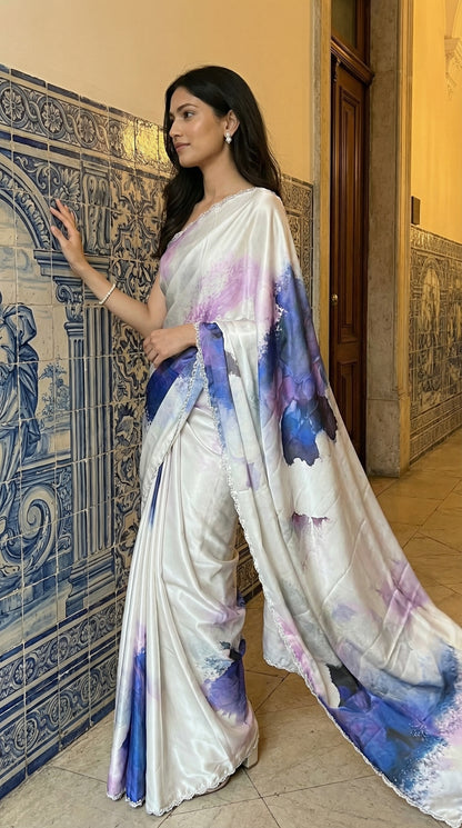 White satin georgette saree with abstract blue brushstroke print and scalloped border, standing against a decorative wall.