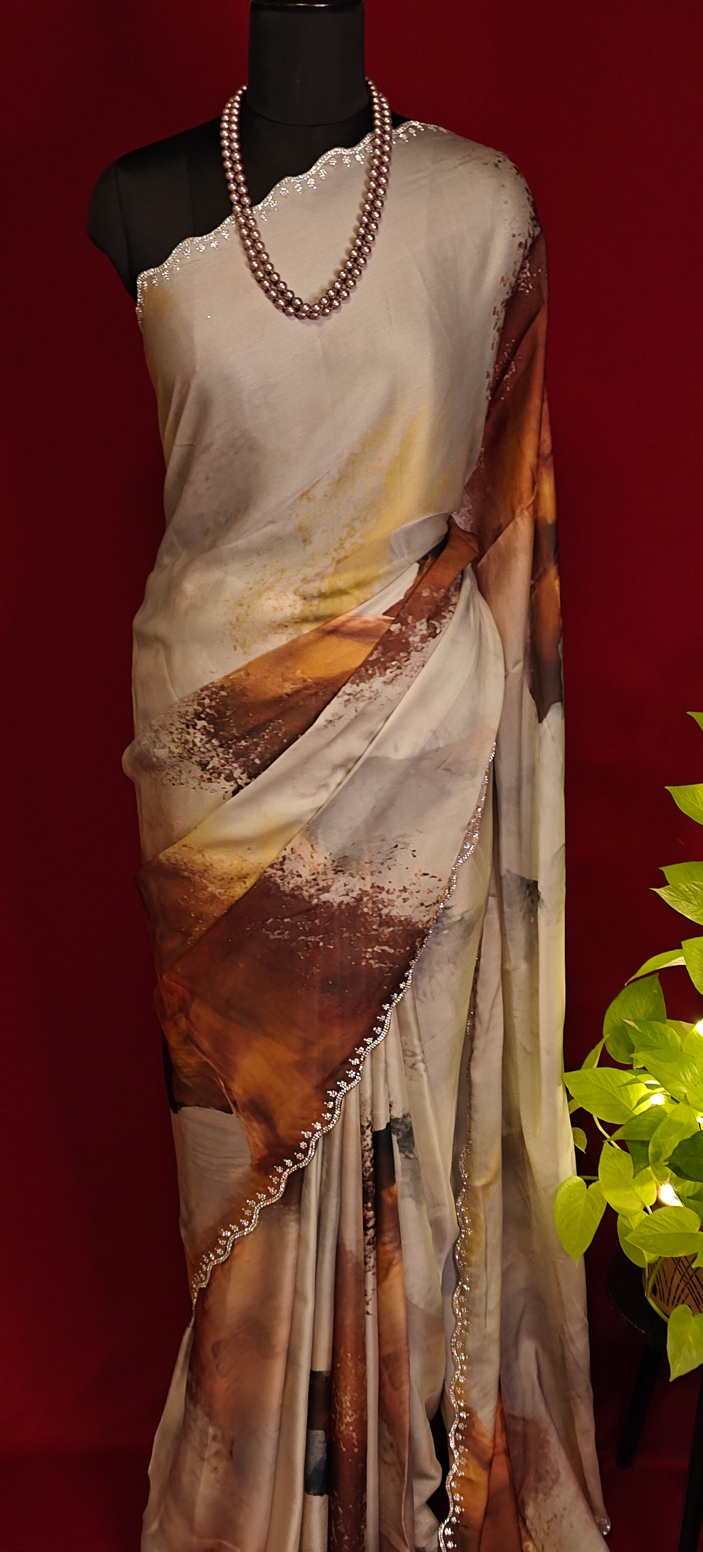 White satin georgette saree with abstract brown brushstroke print and scalloped border on a mannequin with a red background and a plant to the side.