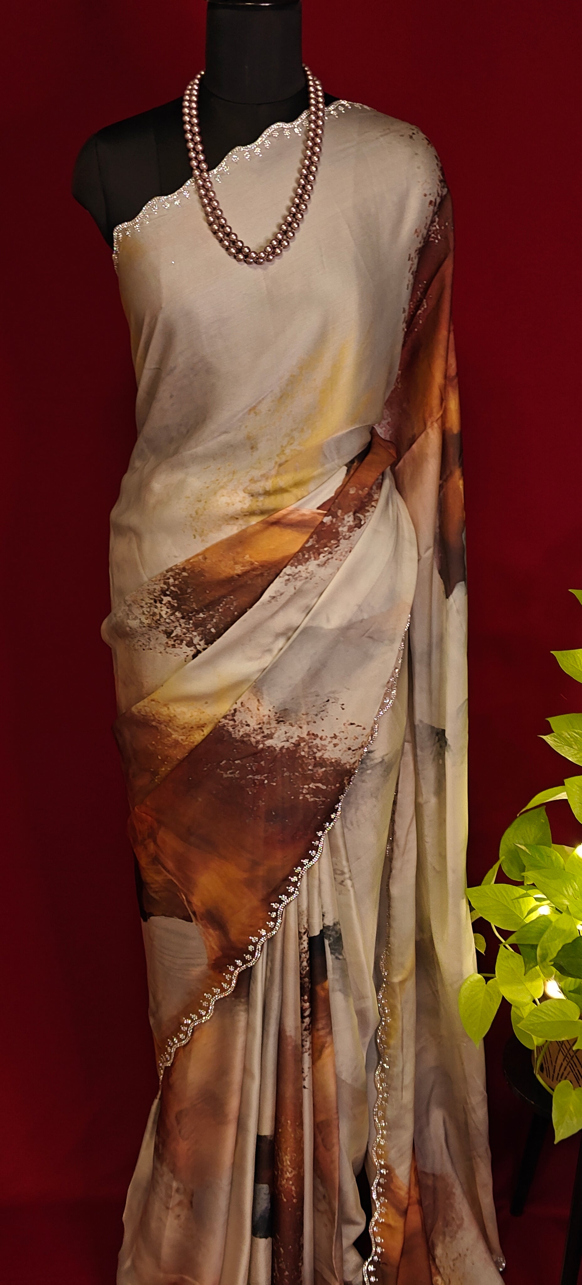 White satin georgette saree with abstract brown brushstroke print and scalloped border on a mannequin with a red background and a plant to the side.