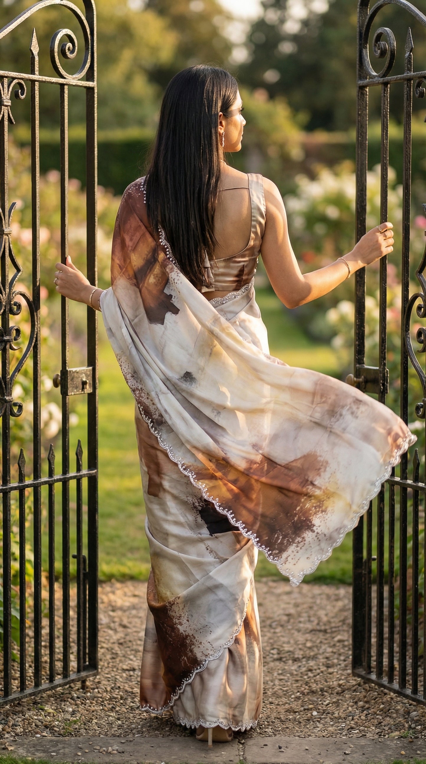 White satin georgette saree with abstract brown brushstroke print and scalloped border, standing in front of an ornate gate with a garden background.
