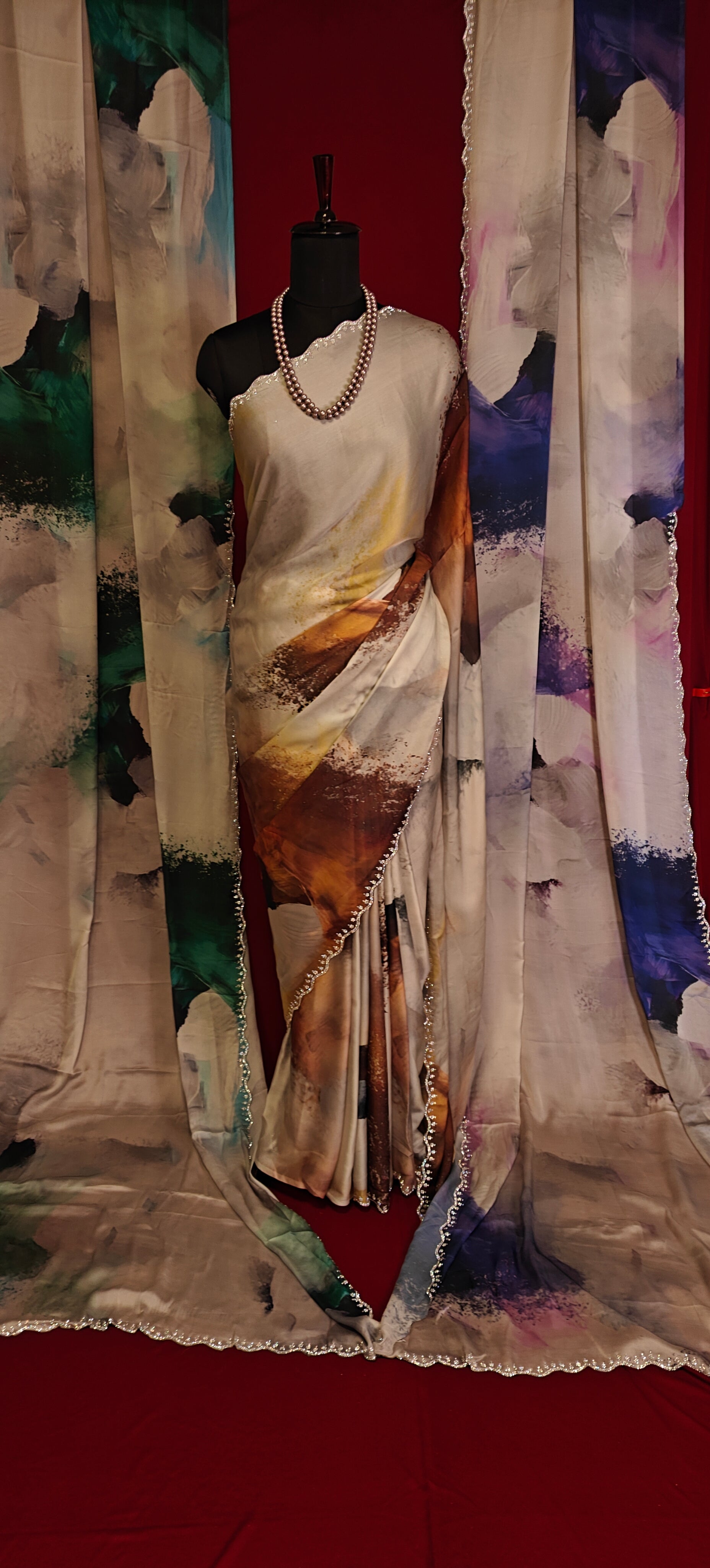 White satin georgette saree with abstract brown brushstroke print and scalloped border on a mannequin along with similar saree in blue and green hanging side by side with a red background.