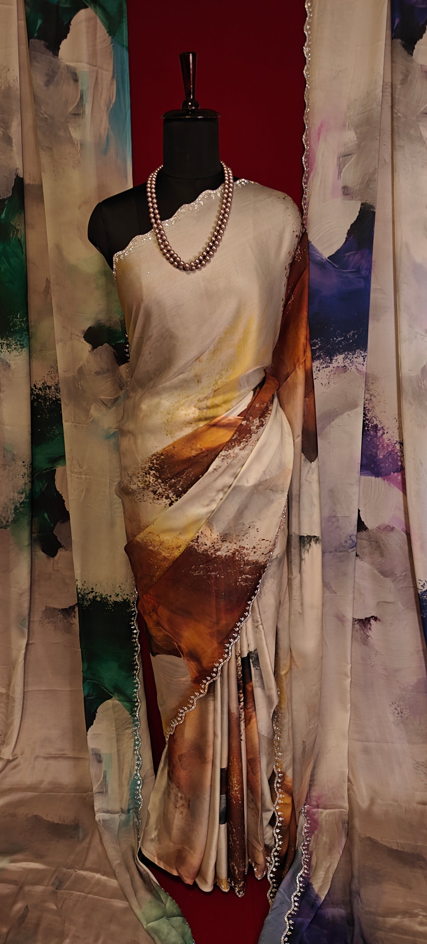 White satin georgette saree with abstract brown brushstroke print and scalloped border on a mannequin along with similar saree in blue and green hanging side by side with a red background.